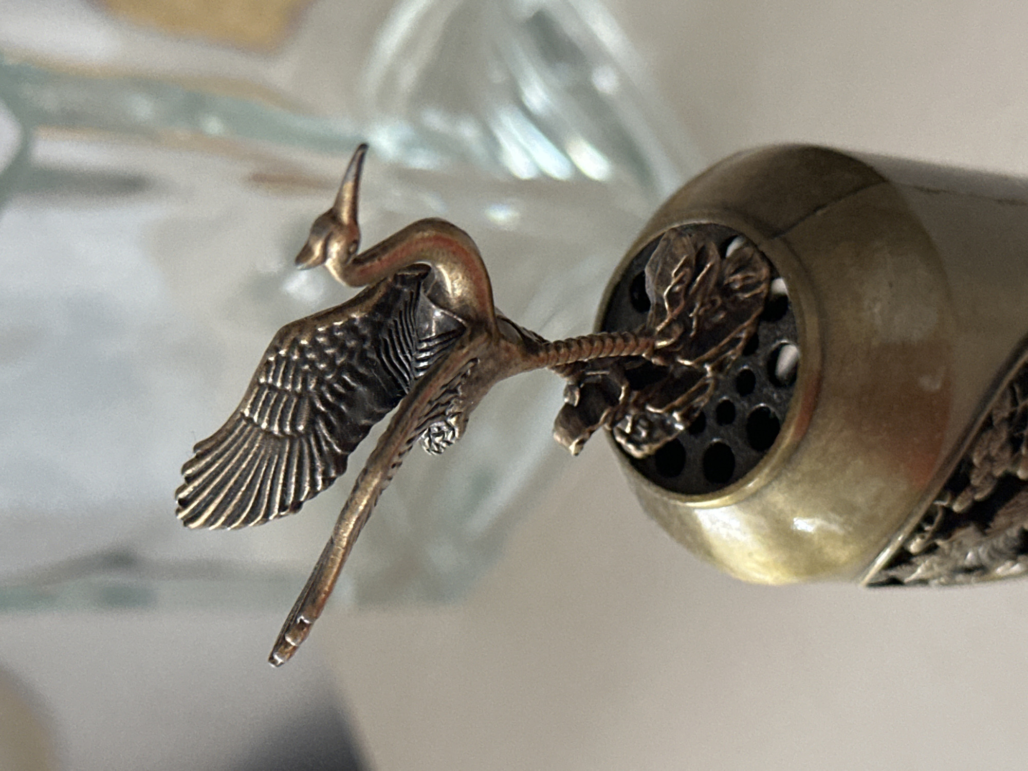 A close-up shot of a brass-colored ornament on top of a cylindrical object. The ornament is a detailed sculpture of a bird, possibly a crane, with its wings spread and its beak pointed forward. The bird is perched on a textured base that is part of the cylindrical object. The cylindrical object appears to be made of metal and has a decorative pattern around its lower section. The background is blurred, showing hints of glass objects and a neutral-colored surface. There is no information about the city of Nevinnomyssk, Russia, in the image.