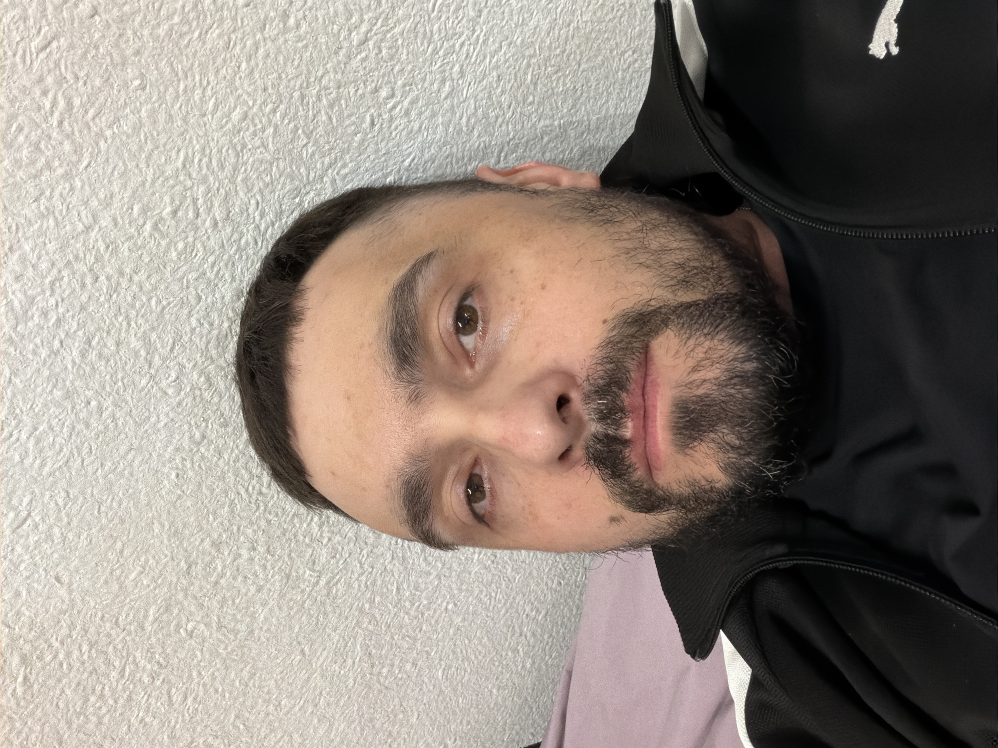 A close-up headshot of a man with short, dark hair and a dark beard. He has light skin, brown eyes, and is wearing a black jacket. The background is a textured white wall. The image provides no information to suggest a location in the city of Kovel, Ukraine.