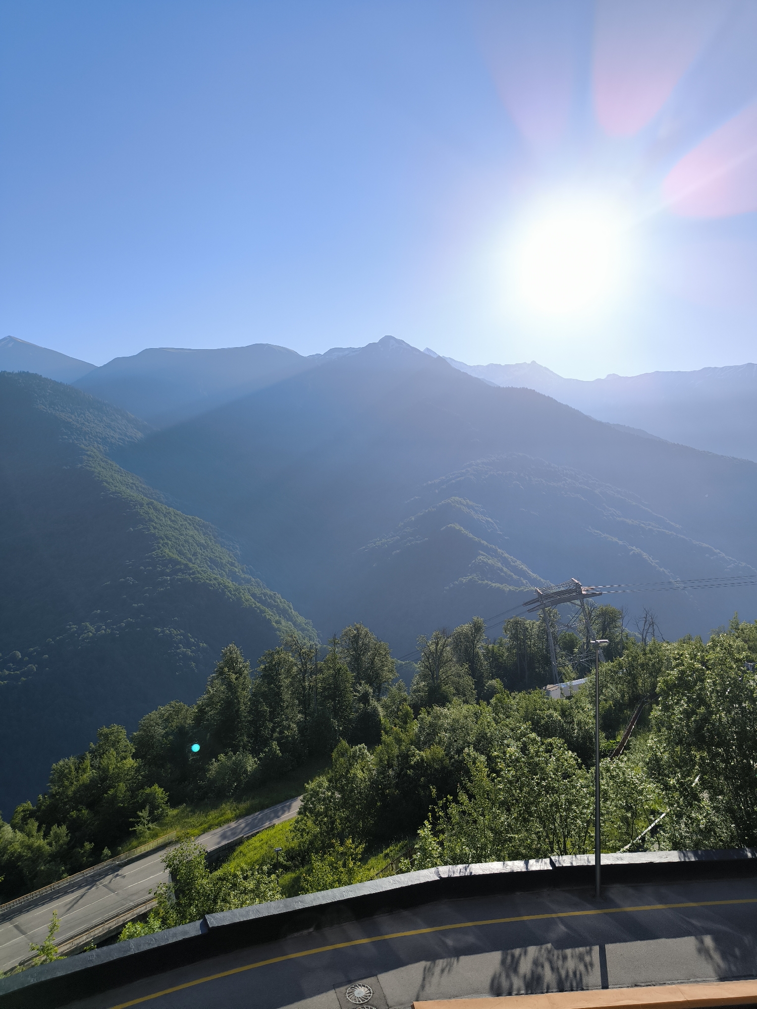 In the city of Sochi, Russia, a photograph captures a majestic mountain landscape bathed in the bright sun. The sky is a clear, vibrant blue, with the sun positioned high and to the right, casting a lens flare across the upper portion of the image. Layers of mountains recede into the distance, their slopes covered in lush green forests. The foreground features a winding road with yellow markings, bordered by a dark railing. Tall trees with dense foliage line the side of the road, and a light pole stands near the edge. A cable car pylon with cables stretching across the frame is visible amidst the trees and mountains, suggesting a recreational area. The scene evokes a sense of natural beauty and peacefulness, characteristic of Sochi's mountainous regions.