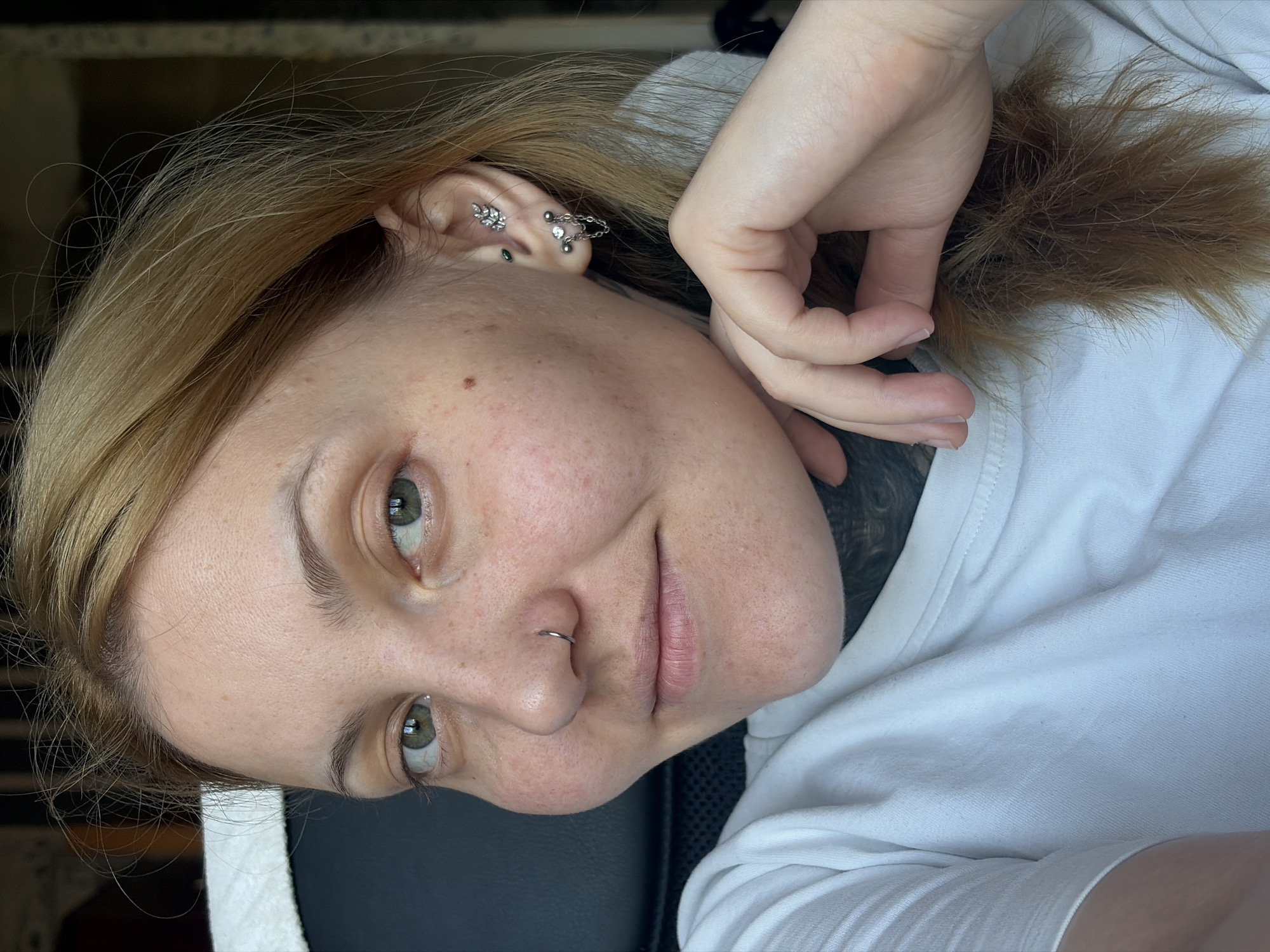 A close-up, portrait-style image shows a person with fair skin and light brown and blonde hair. The individual has light green eyes, freckles on their face, and a silver nose ring. The person is wearing multiple earrings in their left ear, including a silver leaf-shaped earring, a chain earring, and other studs. The person is also wearing a white t-shirt. Part of a dark tattoo is visible on the individual’s neck. The background shows a dark surface and a glimpse of a light-colored wall. The image does not provide enough information to confirm the location as Chaikovsky, Russia.