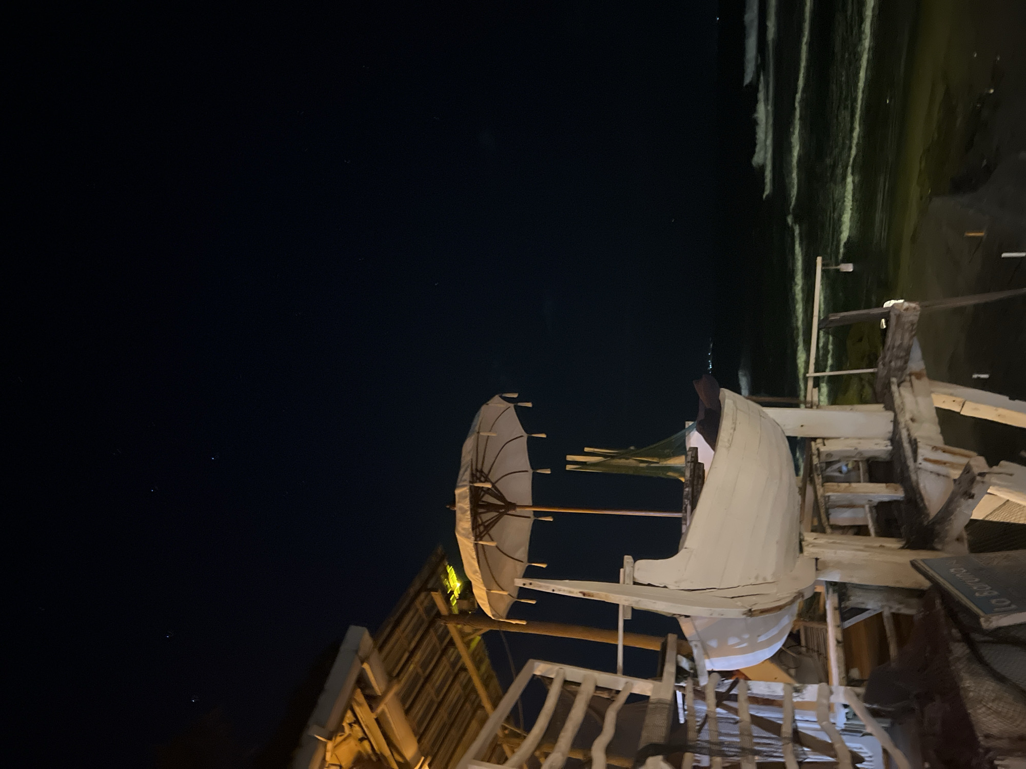 The image shows a night scene, likely taken in North Kuta, Indonesia, where artificial structures are illuminated against a dark sky. In the foreground, a white, somewhat dilapidated, wooden structure is visible. A partially open umbrella is attached to the structure, along with other fragmented wooden elements that suggest a nautical theme. The structure seems to be on a raised platform, evidenced by the presence of railings and stairs. The scene is backlit. In the distance, a dark beach is visible along with some waves.
