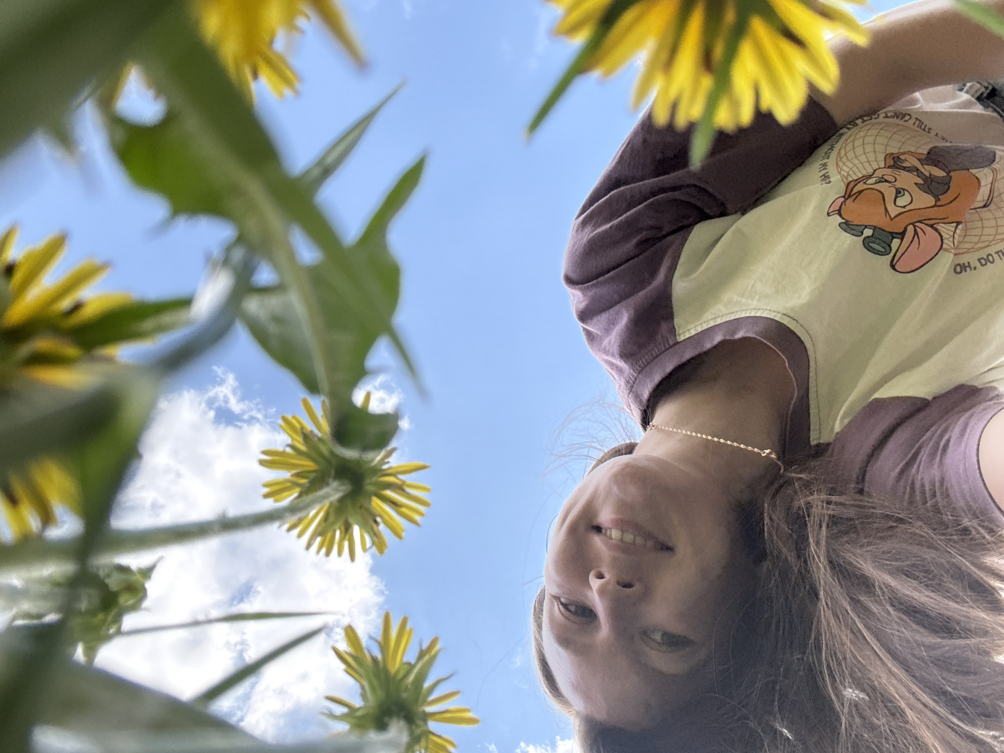 A low-angle, upward-facing shot shows a woman in the foreground, with her head partially obscuring a bright, clear blue sky dotted with fluffy white clouds. Yellow dandelion flowers with green stems and leaves surround the top and sides of the frame, creating a border. The woman is wearing a two-tone shirt with brown sleeves and a lighter-colored body, featuring a cartoon character on the front. She has fair skin, light brown hair, and is smiling. A thin gold necklace is visible around her neck. The location is assumed to be Italmas, Russia.