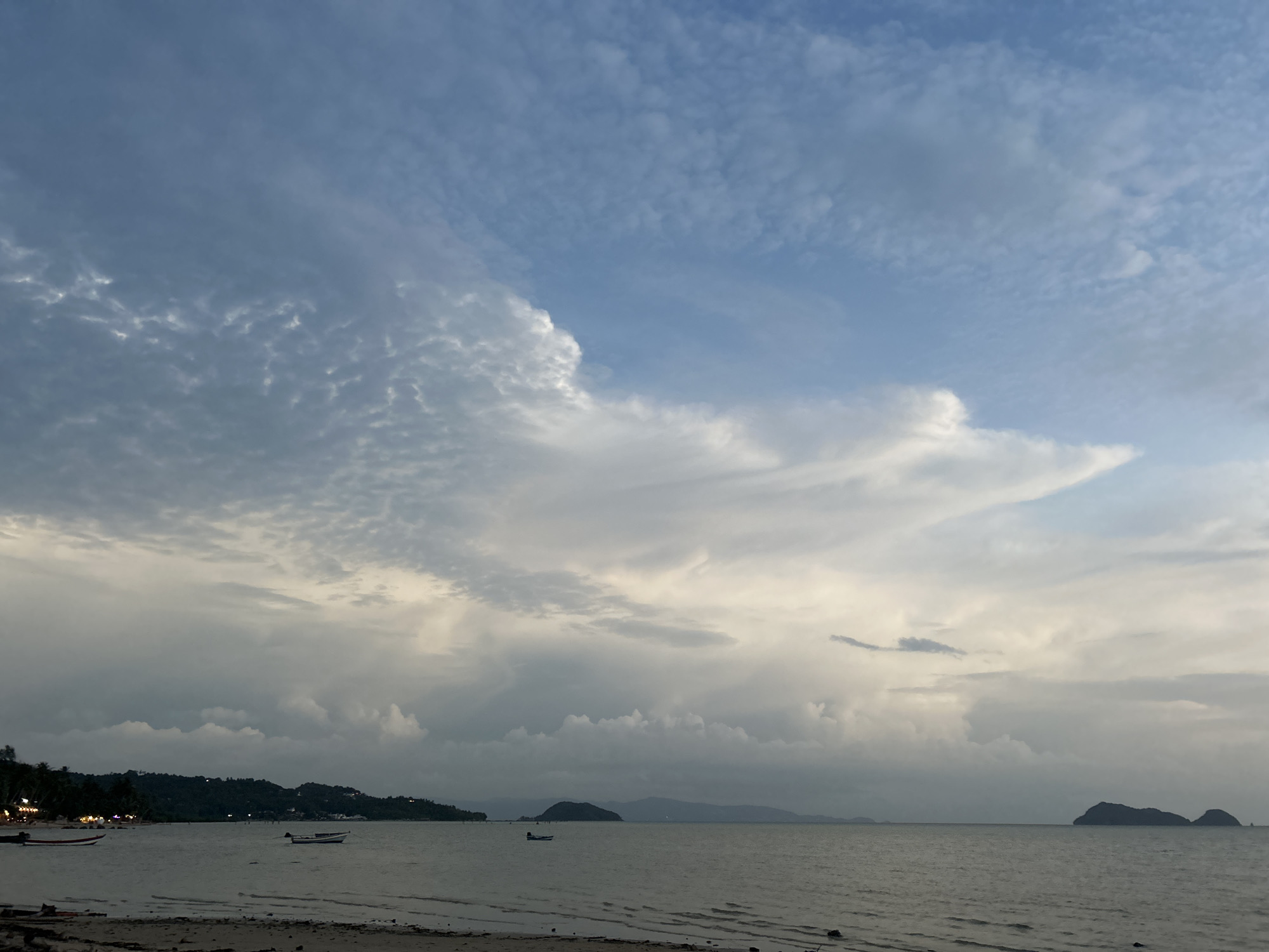 The image depicts a coastal scene at dusk or early morning in Ko Pha-ngan District, Thailand. The sky is a mix of pale blue and white clouds, with some areas appearing overcast. The sea is calm, with gentle waves lapping at the shore. In the distance, a dark, tree-covered island can be seen, with smaller islands dotting the horizon. A few small boats are anchored in the water, and some lights are visible along the coastline, suggesting a village or resort area. The beach in the foreground is sandy and appears to be mostly deserted. The overall impression is one of tranquility and natural beauty.