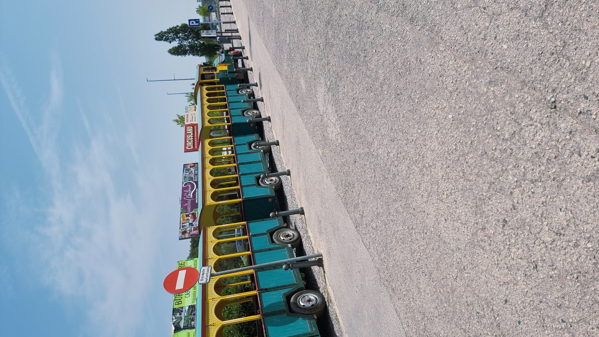 The image depicts a stationary tourist train, painted teal and yellow, parked along a paved pathway. The train consists of multiple open-air carriages. In the background, there are several billboards advertising local attractions, including "BURE CASTELLANO TERRASSA AMB", "JARDINS LAXARTANA", and "CIRCUSLAND". A red and white circular "No Entry" sign is visible near the front of the train, with a smaller sign below it reading "Excepte autoritzats". The environment appears to be outdoors, under a clear blue sky with some wispy clouds. The ground is covered in asphalt and gravel. The presence of tourist-oriented signage and the train itself suggest this is a location catering to visitors, possibly near a park or attraction in or around Besalú, Spain. No people are visible in the scene.