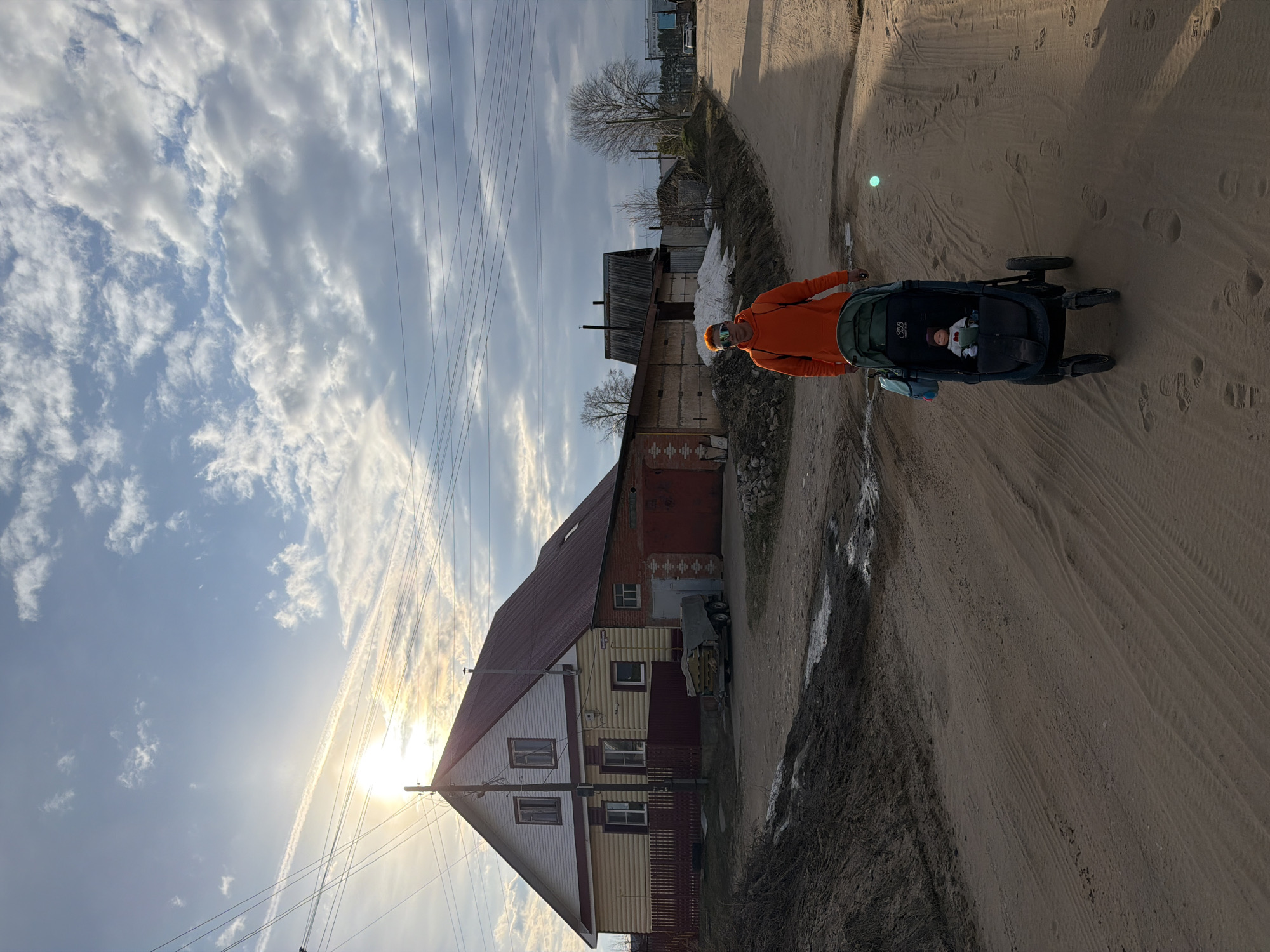 The image depicts a person pushing a baby in a stroller along a sandy dirt road, likely in Chaikovsky, Russia. The scene is captured during late afternoon or early morning, with the bright sun low in the sky, casting a warm glow and creating lens flare on the left side.
A person wearing a vibrant orange hoodie and reflective sunglasses is pushing a dark-colored baby stroller. Inside the stroller, a baby is visible, wearing a hat. The stroller has the brand names "SJS" and "CITY MINI" printed on it. The road is unpaved, showing clear tire tracks and footprints, with some muddy patches and remnants of snow or ice along the edges, suggesting early spring or late autumn.
In the mid-ground, to the left, a two-story house stands prominently. It features a dark red gabled roof and a facade combining yellow and white siding. A utility pole with numerous power lines runs past the house and extends across the sky. Adjacent to the house, a vehicle is partially visible under a cover. Further in the background, other buildings and bare trees can be seen under a partly cloudy sky, with long, wispy clouds and what appear to be airplane contrails. The setting evokes a peaceful, semi-rural environment.