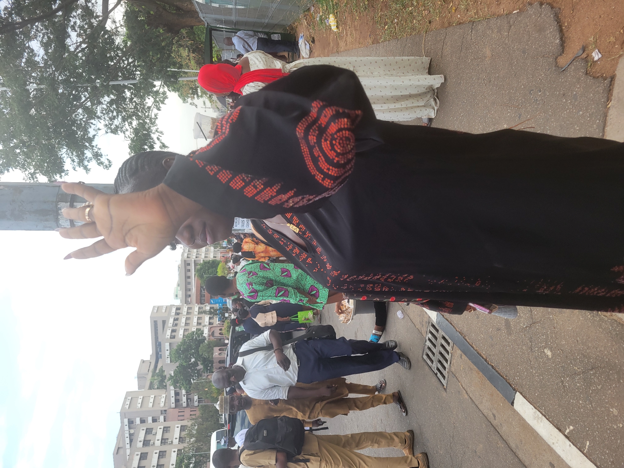 The scene depicts a crowd of people standing and walking on a paved street in Abuja, Nigeria. Buildings are visible in the background. A person with a hand raised toward the camera dominates the foreground. Other people are wearing various clothing, including a black robe with orange patterned trim, a red headscarf, and patterned green fabric. Some people are wearing uniforms.
