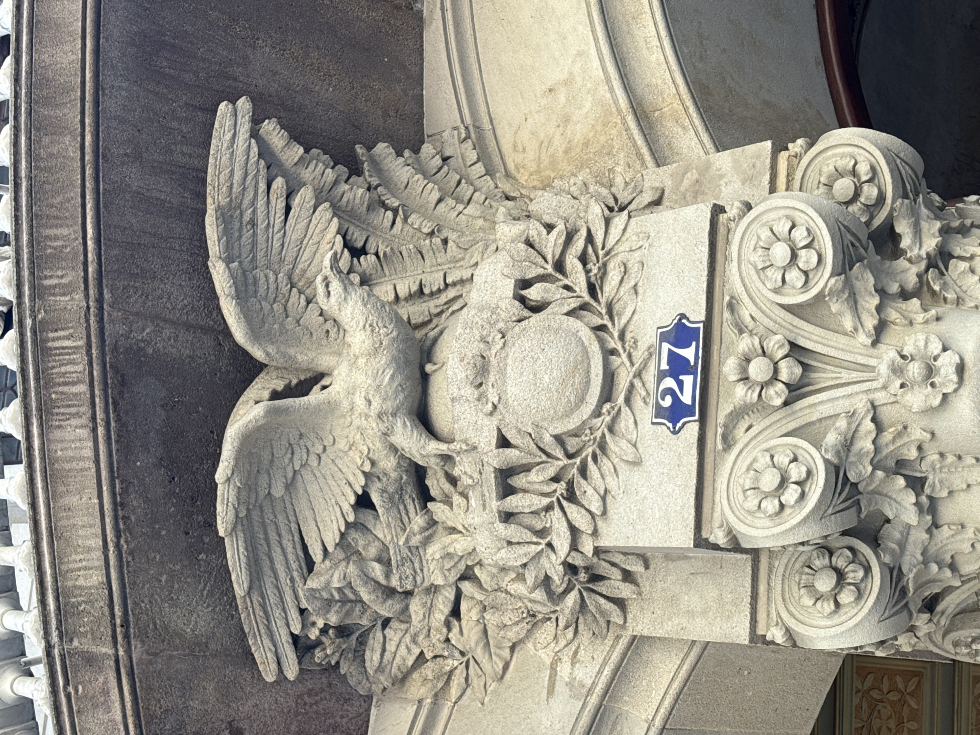 The image displays a close-up of an ornate architectural detail, likely a building facade in Barcelona, Spain. The primary subject is a stone carving featuring a stylized eagle with outstretched wings, holding a spherical object in its talons. The eagle is surrounded by carved foliage, possibly laurel leaves, adding to the classical and elaborate aesthetic. To the right of the eagle carving, a small blue and white plaque is visible, bearing the number "27" in white numerals. This plaque indicates a building number. The stonework is highly detailed, with individual feathers of the eagle, the veins of the leaves, and floral motifs carved with precision. The color of the stone is a light grey, weathered by time. The background is a mix of darker stone or plaster, creating a contrast with the lighter sculpture. The lighting suggests it is daytime, possibly overcast given the diffused light. The overall impression is one of historical architecture and artistic craftsmanship. There is no indication of people or any specific event taking place.