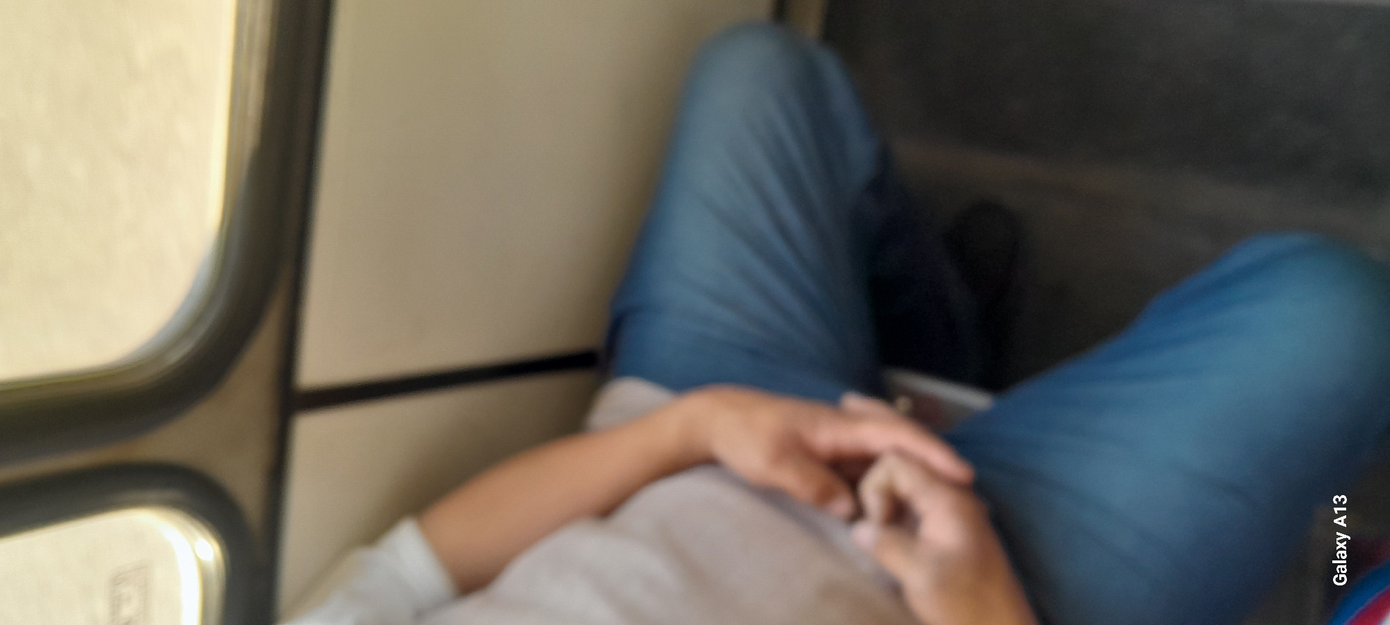 The image shows a person wearing a light-colored t-shirt and blue jeans, sitting on a seat with their legs crossed. The person's hands are clasped together in their lap. The background consists of a window with a view of what appears to be a light-colored wall. There is a dark floor visible beneath the person's legs. The scene is indoors, possibly on public transportation, given the window and seating arrangement. The lighting suggests it is daytime. The bottom left corner of the image displays the text "Galaxy A13," indicating the device used to capture the photograph. No specific location in the city of Angren, Uzbekistan can be determined from the visual information present in the image.