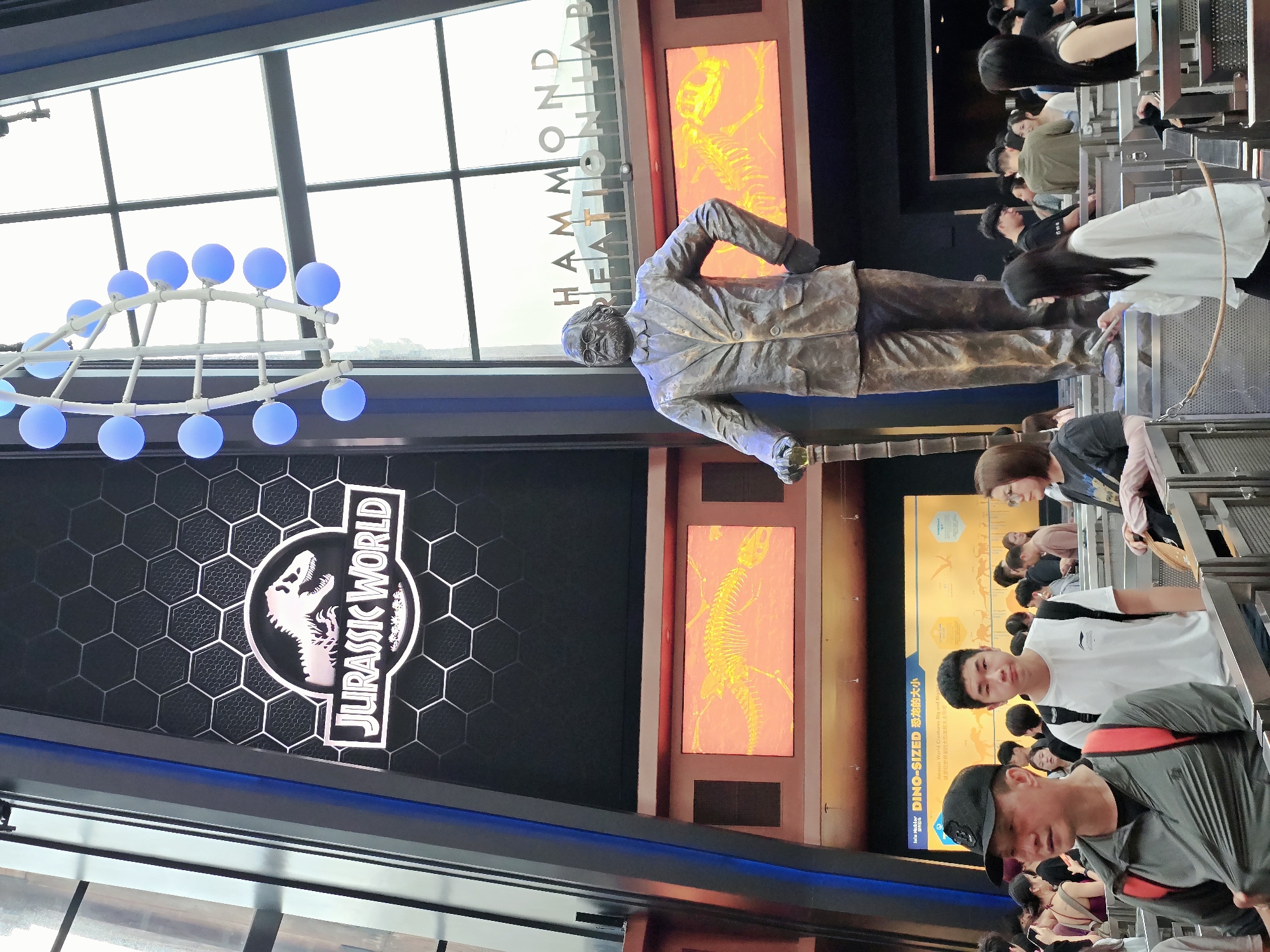 The image captures the entrance area of Jurassic World, located in Universal Beijing Resort in Tongzhou District, China. Dominating the scene is a large bronze statue of John Hammond, the fictional founder of Jurassic Park, holding a cane. Behind him, a large screen displays the Jurassic World logo, featuring a dinosaur skeleton, set against a hexagonal pattern. Further down, another screen shows a yellow dinosaur skeleton on an orange background. The environment is grand and themed, with architectural elements suggestive of a theme park. A diverse crowd of visitors is visible, some queuing and others walking, indicating an active attraction. The lighting is a mix of ambient light from the screens and overhead fixtures, with a decorative chandelier featuring blue spherical lights hanging from the ceiling. The overall impression is that of a lively, immersive tourist destination.