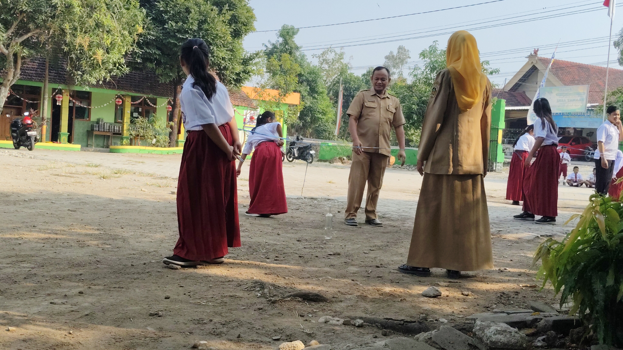 In Banjarsari, Indonesia, a group of schoolchildren, mostly girls in white shirts and maroon skirts, stand in a dusty outdoor area, possibly a schoolyard. A male teacher in a tan uniform stands in the middle, holding a string taut, seemingly engaged in an activity with the students. Some of the girls are positioned in front of him, looking towards him or the ground. Another female figure, wearing a tan uniform and a yellow hijab, stands facing away from the camera. The background shows a green building with red and white decorations, trees, a parked motorcycle, and other students sitting in the distance. The overall scene appears to be a school event or activity taking place outdoors under daylight.