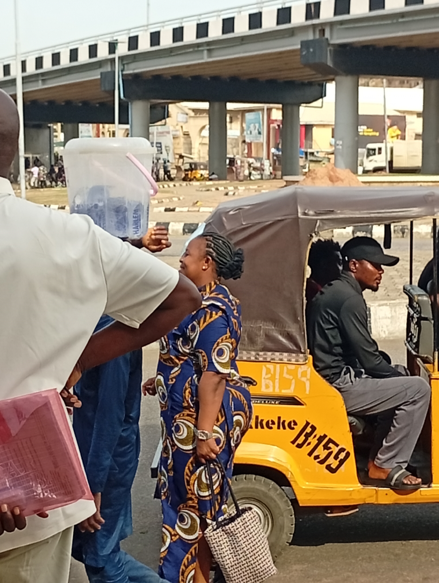 The image depicts a street scene in Lokoja, Nigeria, near a concrete overpass. A yellow "keke" (auto-rickshaw) labeled "B159" is a prominent subject. A woman in a blue patterned dress with a woven handbag is interacting with a man selling water from a bucket. The man is carrying a bucket labeled "HARLEM". Another man in a black cap is driving the keke. Other people and vehicles are in the background.