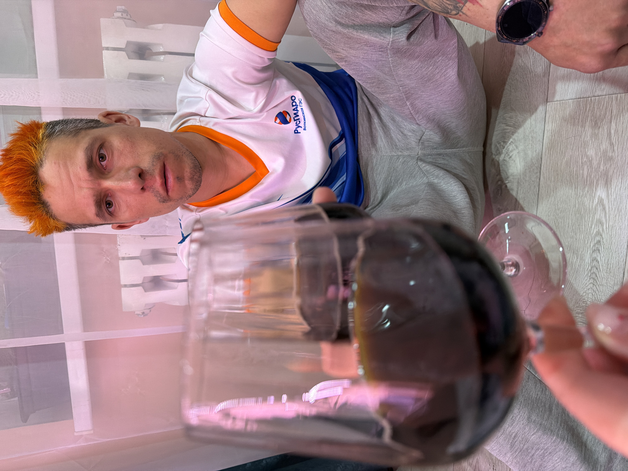 The image depicts a man with orange hair, wearing a white t-shirt with blue accents and the "РусГидро" logo, sitting on a light-colored floor. He has a digital watch on his wrist and a tattoo on his arm. In the foreground, there is a glass of dark liquid, possibly wine, partially obscuring the view. Behind the man is a radiator. The location is Chaikovsky, Russia.