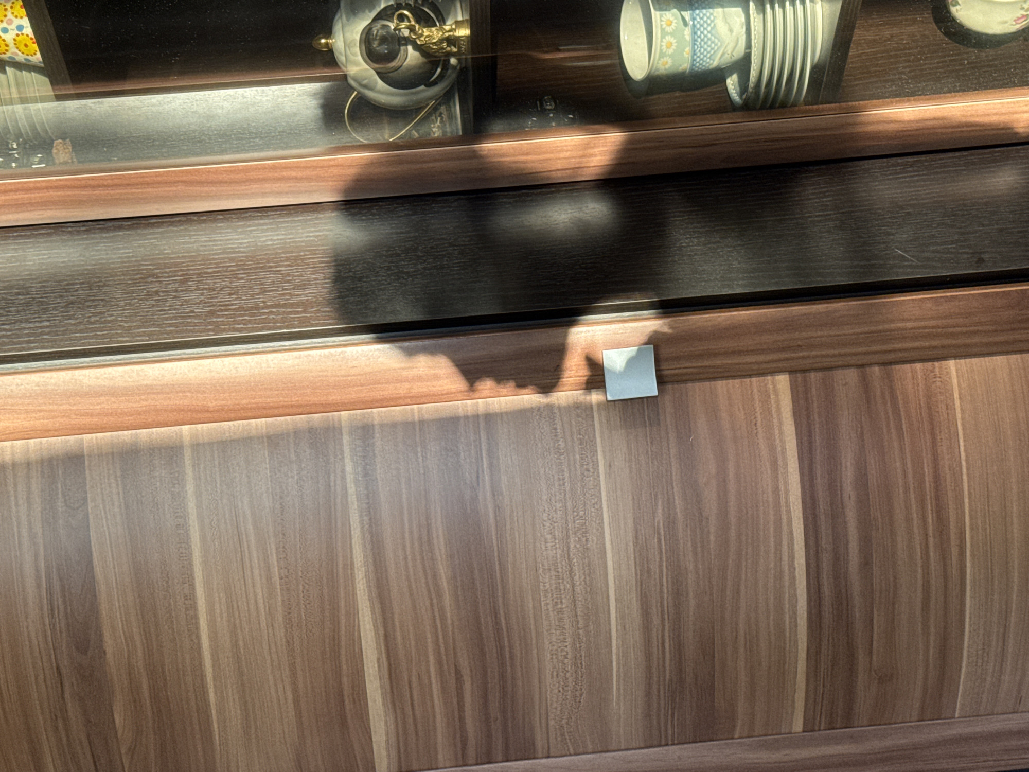 A low-angle, close-up photograph shows a portion of a cabinet, with a shelf exhibiting teacups and decorative items under a glass display. The cabinet has multiple horizontal sections with a brown wood grain pattern, the bottom section has vertical grains. On the horizontal section with the vertical wood grains, there is a square-shaped piece of white paper. The photograph appears to be taken indoors under natural light. The location and additional details of the city Izhevsk, Russia, are not discernible from the provided image.