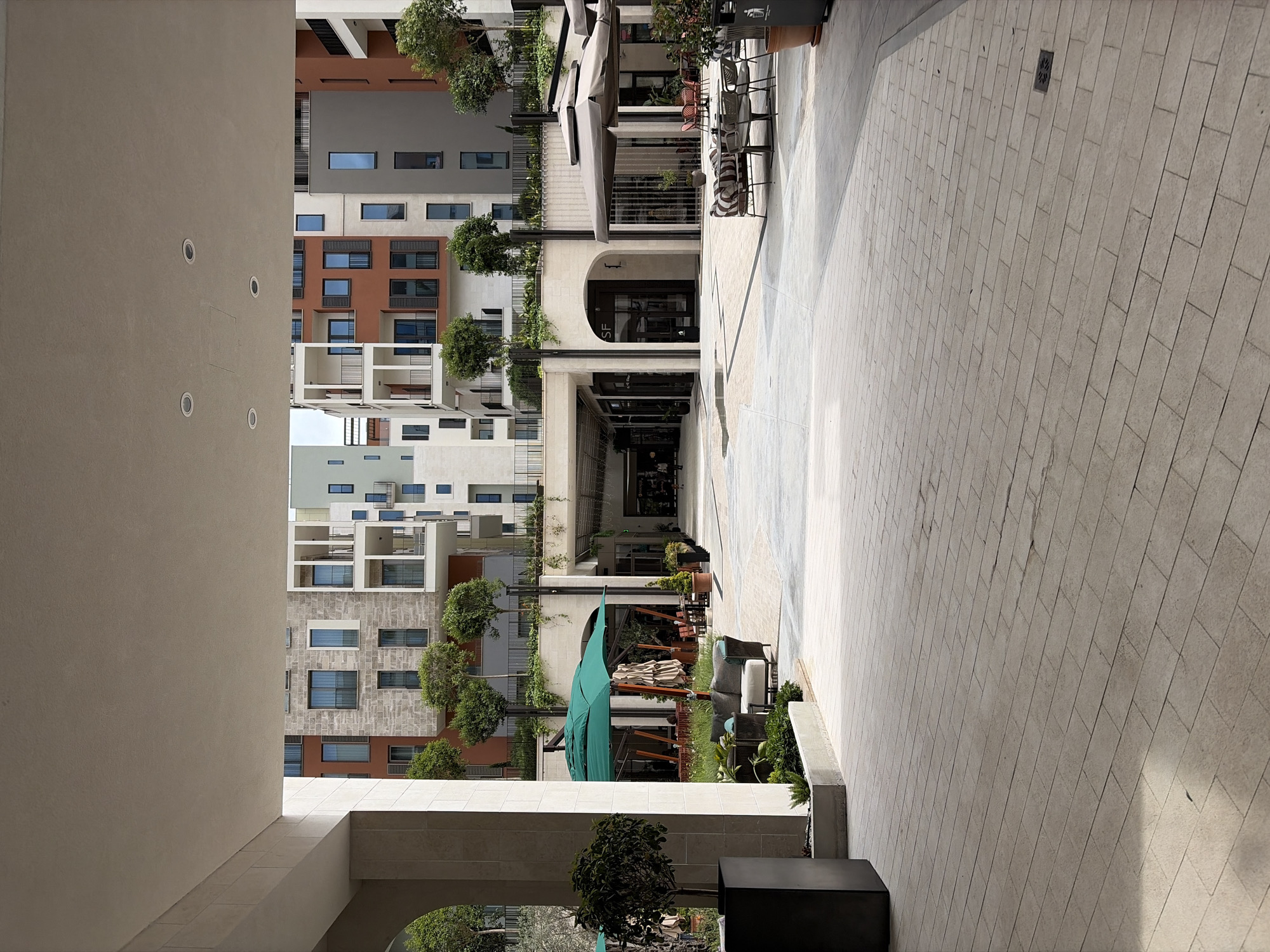 The image shows a pedestrian street in Porto Montenegro, Tivat, Montenegro. The street is paved with light-colored rectangular blocks. Modern buildings with multi-colored facades (white, brown, gray, stone) line the left side of the street. Some buildings have balconies with metal railings. Trees are planted along the buildings. Shops and restaurants are along the street, with outdoor seating areas featuring tables, chairs, and umbrellas. A large green umbrella is visible in one of the seating areas. There are no people clearly visible. The photo is taken from under a covered walkway.