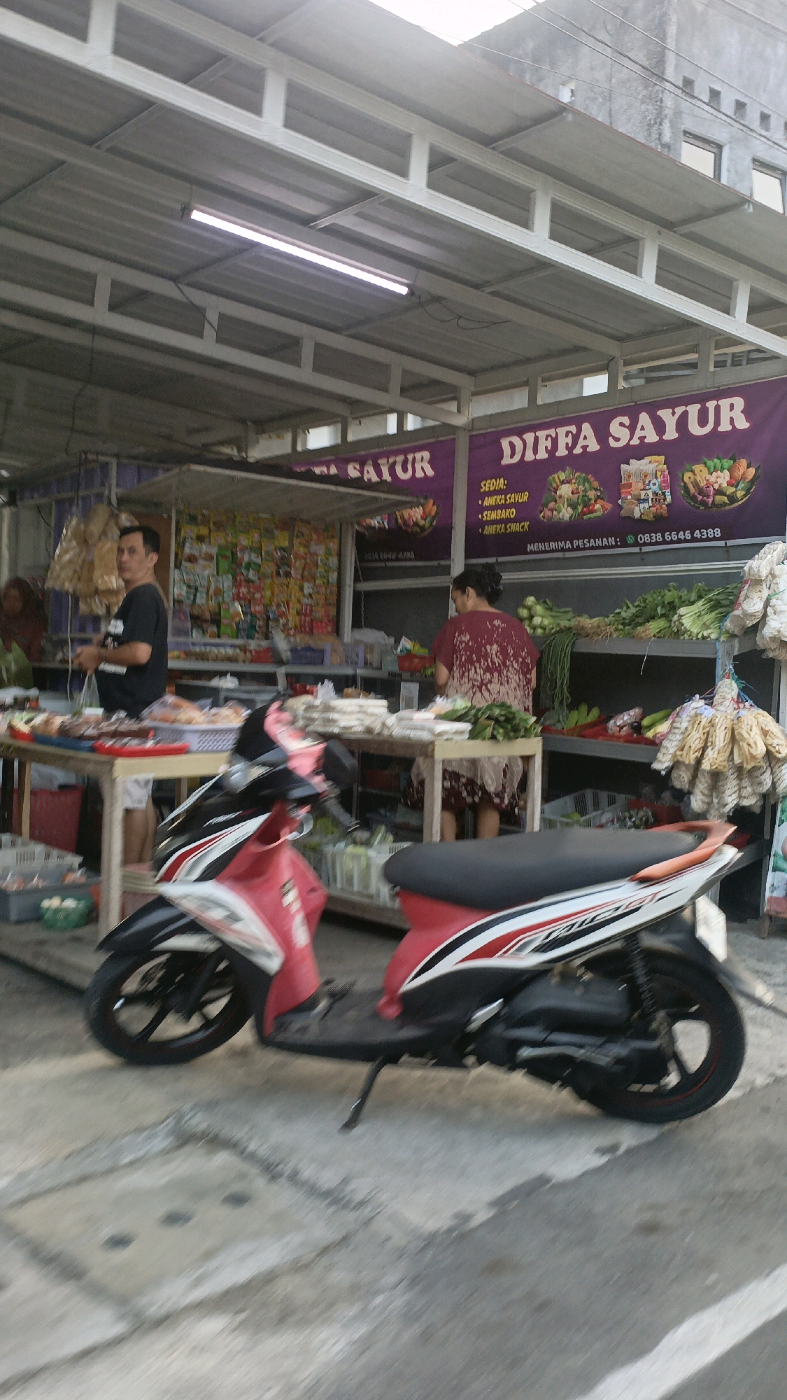 This is a medium-shot, eye-level, vertical, outdoor photograph taken in Salatiga, Indonesia. The scene depicts a market stall with a woman and a man standing. The stall is under a white metal awning, with a purple banner above that reads "DIFFA SAYUR" and lists "SEDIA: ANEKA SAVUR, SEMBAKO, ANEKA SNACK." A red and black scooter is parked in the foreground, partially obscuring the view of the stall. The woman, wearing a maroon shirt with white patterns, is facing away from the camera, arranging produce on a table. The man, wearing a black t-shirt and shorts, is standing to the left, facing the camera, holding a plastic bag. Tables laden with various goods, including vegetables, snacks, and other items, are visible behind them. The ground is paved with concrete, and a square drain cover is visible in the lower portion of the frame. The overall lighting suggests it is daytime.