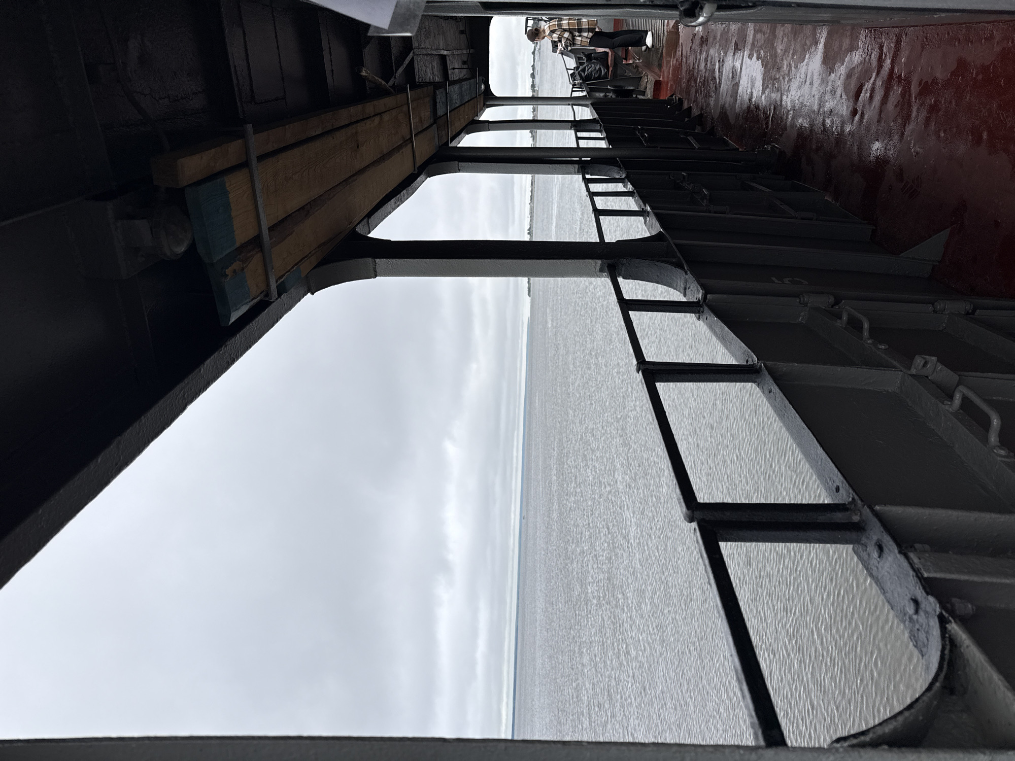The image depicts a view from the deck of a vessel, likely a ferry, on a cloudy day. The foreground features the dark, metal railing and deck of the boat, with a portion of the red-painted floor appearing wet. In the middle ground, a person wearing a plaid shirt and dark pants stands near a black bag and a bench, looking out towards the water. The water itself is grey and calm, stretching to the horizon under a heavily overcast sky. The background shows a faint outline of land on the distant horizon. The structure of the boat's upper deck is visible, with wooden planks and structural supports. The scene is consistent with the Volga River environment in Kozmodemyansk, Russia, where such ferries operate.