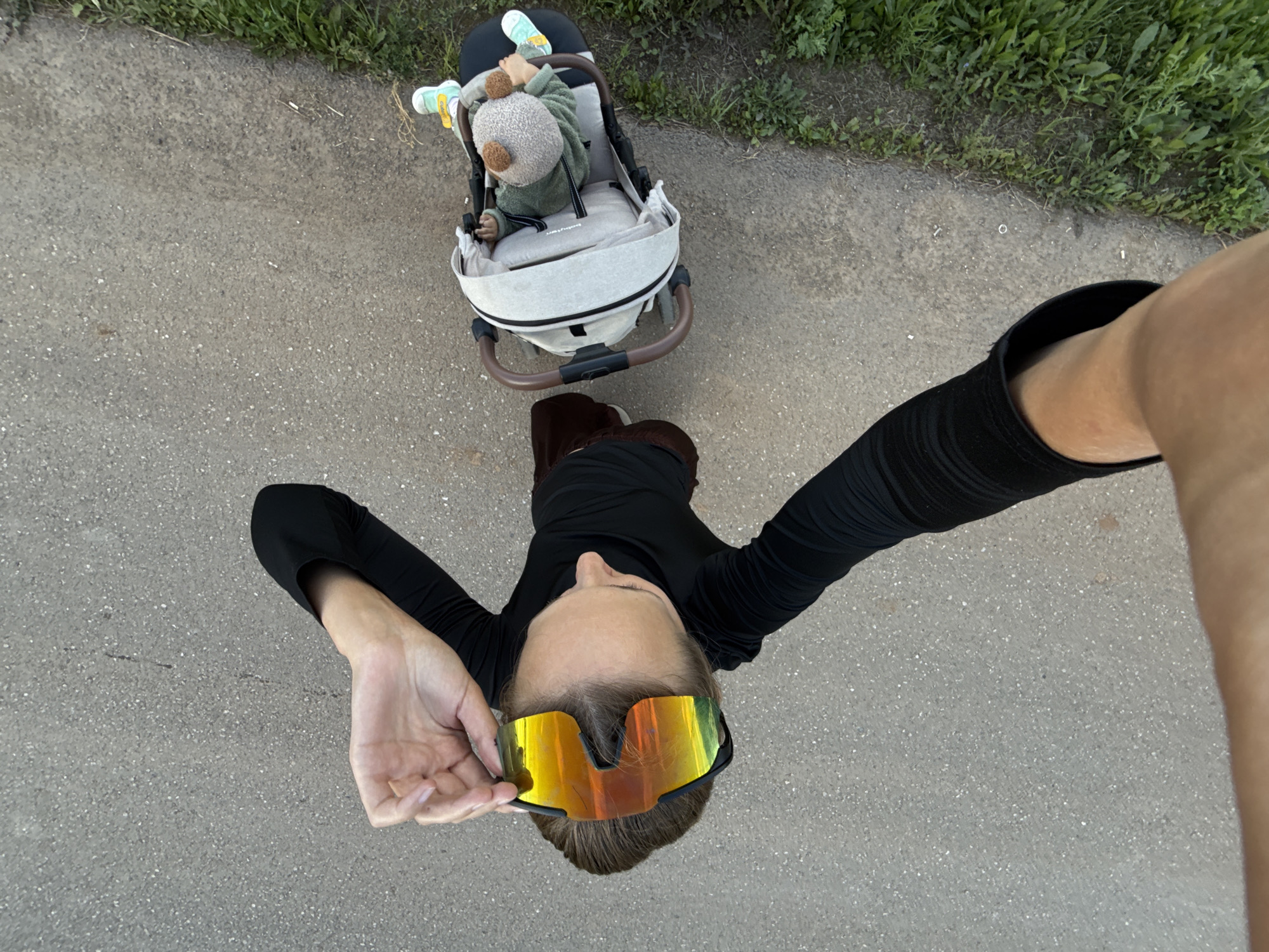 In a high-angle shot on a paved pathway adjacent to a grassy area, a person is pushing a stroller. The person is wearing a black long-sleeved shirt and brown pants, with large, reflective goggles on their face. Their hands are near the goggles, suggesting they are adjusting them. The stroller is light-colored with a brown frame and contains a baby dressed in a green outfit and a beige hat with bear ears. The baby's feet are visible, wearing light blue and yellow shoes. The environment appears to be outdoors in a park or a similar urban outdoor setting, consistent with a location in the city of Podshivalovo, Russia.