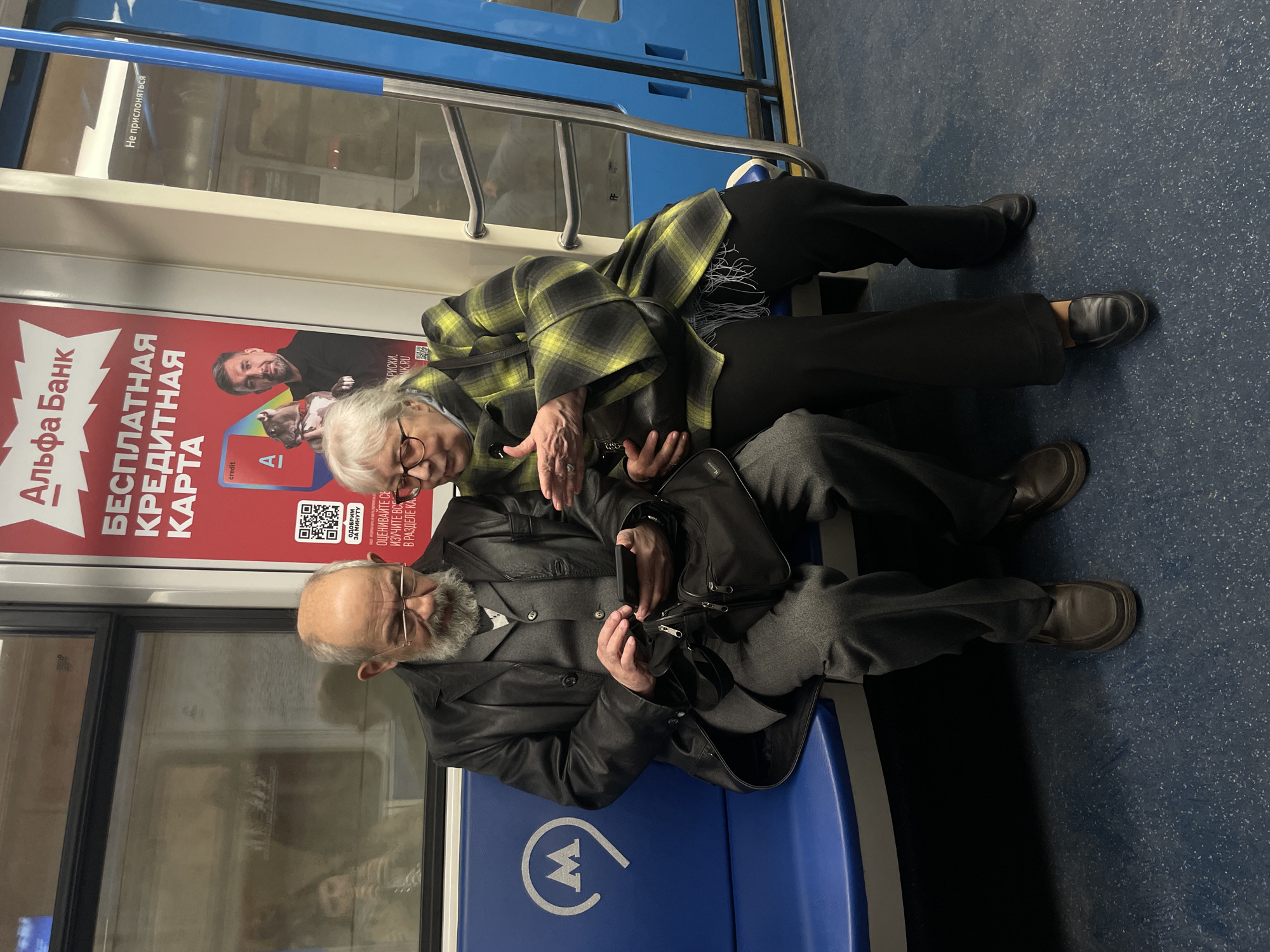 Two elderly individuals, a woman and a man, are seated on blue benches inside a metro train car. The image is taken from a slightly elevated perspective, looking down at them. The woman, with light-colored hair and glasses, wears a distinct yellow and black plaid coat over dark clothing and has a small black handbag on her lap. She is leaning towards the man, gesturing with her right hand, seemingly pointing at or discussing something. The man, with a beard and glasses, is dressed in a dark leather jacket over a gray suit jacket and a white shirt. He is holding a dark-colored device, possibly a smartphone or tablet, in his hands, focused on its screen. A black messenger bag rests on his lap.
The setting is a public transport environment, clearly identifiable as a subway or metro train due to the typical blue seating, the windows with reflections, and a prominent white "M" logo (characteristic of the Moscow Metro) on the blue seat backing. The floor is a dark blue with a textured, speckled pattern.
A large red advertising poster is visible on the wall behind the woman. The activity suggests a quiet interaction between the two, perhaps reviewing something on the man's device. The lighting within the train car appears consistent and bright, indicating an indoor setting regardless of the time of day.
Visible text in the image includes:
* On the red poster: "Альфа Банк" (Alfa Bank), "БЕСПЛАТНАЯ КРЕДИТНАЯ КАРТА" (FREE CREDIT CARD), "ОДОБРИМ ЗА МИНУТУ" (Approved in a minute), and partially visible phrases like "credit A", "ОЦЕНИВАЙТЕ СЕ / ИЗУЧИТЕ ВСЕ / В РАЗДЕЛЕ КА" (Evaluate / Learn everything / In the section), and what appears to be a URL "КРИСКИ.NK.RU". A QR code is also present.
* On the window: "Не прислоняться" (Do not lean / Do not touch).