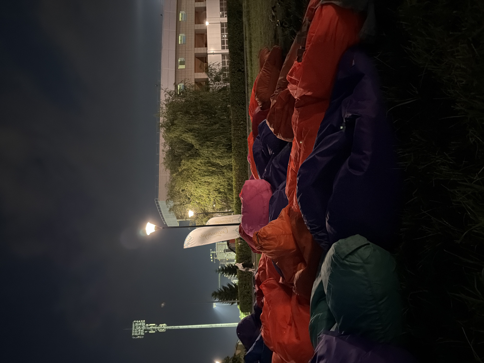 In an outdoor urban setting at night, a large cluster of colorful beanbag chairs fills the foreground, creating a vibrant and informal seating area. The beanbags are predominantly in shades of red, orange, purple, and green, some appearing slightly deflated or jumbled together. Beyond the beanbags, a neatly trimmed hedge forms a boundary, separating the seating area from a grassy lawn. In the middle ground, a willow tree with its distinctive drooping branches stands tall, its leaves illuminated by ambient light. Behind the tree, a modern building with a light-colored facade and multiple windows is visible. The building appears to be a stadium or arena, suggested by its scale and the presence of bright lights on a tall, multi-array tower in the background, which is characteristic of sports venues. A streetlamp with a warm glow illuminates part of the scene. A lone figure walks in the distance on the left side of the image, near the tall tower, and a white flag with dark text stands to the right of the streetlamp. The sky above is dark and overcast, with a hint of lighter clouds. The overall impression is an event space set up for an evening gathering or performance, with the beanbags suggesting a relaxed and communal atmosphere.