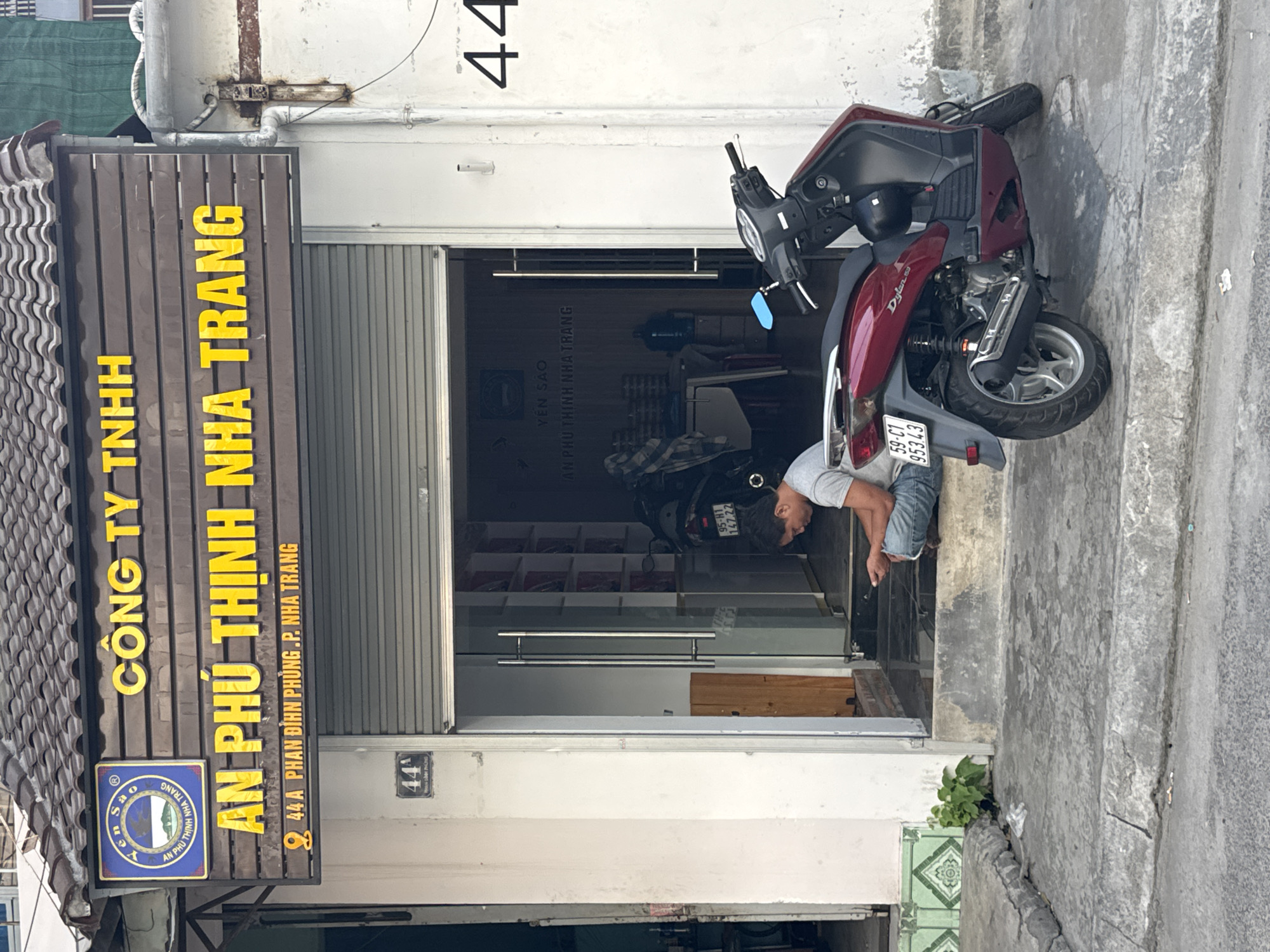 The image shows the storefront of a business named "CÔNG TY TNHH AN PHÚ THỊNH NHA TRANG" located at "44A PHAN ĐÌNH PHÙNG, P. NHA TRANG". The signage is prominent, with large yellow lettering on a dark wooden background. A blue and yellow circular logo is also visible on the left side of the sign.
In front of the storefront, a man is sitting on the steps, looking down at a mobile phone he is holding in his hands. He is wearing a grey t-shirt and blue jeans. A dark red scooter, a Honda Dylan, is parked to the right of the entrance, partially obscuring the doorway. The scooter has a black helmet hanging from the handlebars.
The entrance to the store is open, revealing shelves inside stocked with items, some of which appear to be packaged in red boxes. A large blue water dispenser is also visible inside. The building has a weathered facade with peeling paint. The street in front of the store is paved with concrete, showing cracks and wear. There is some greenery growing from cracks in the sidewalk. The overall scene depicts a typical street view in Nha Trang, Vietnam.