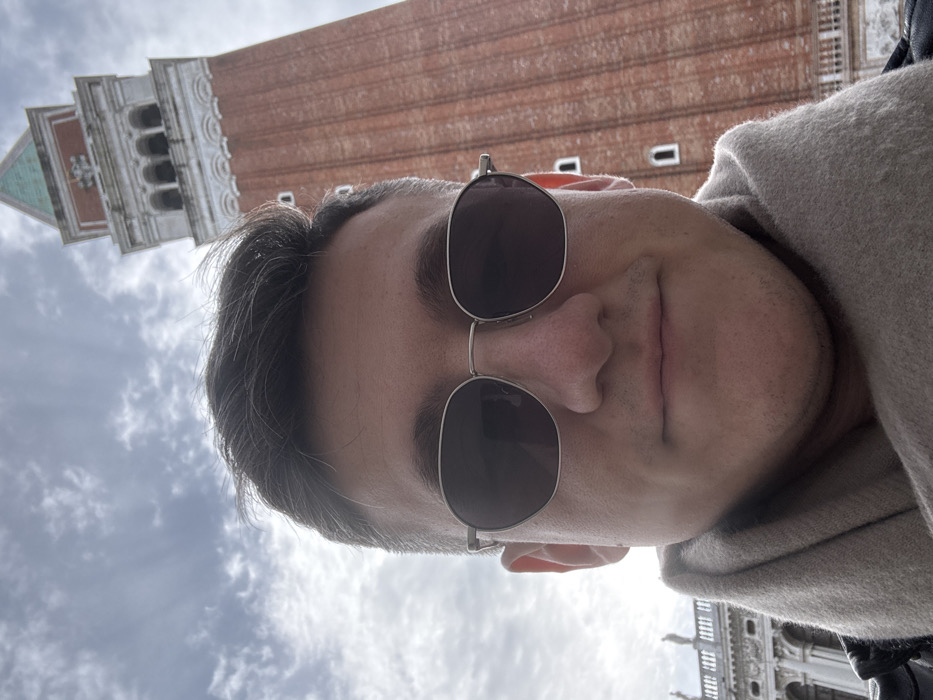 A male individual is depicted in a close-up, low-angle shot, likely a selfie, in St Mark's Square, Venice, Italy. The man occupies the lower and central portion of the frame, wearing dark-lensed, silver-framed sunglasses and a light-beige scarf. He has dark hair and light skin, with a neutral facial expression. Directly behind him, towering into the sky, is St Mark's Campanile (Campanile di San Marco), a prominent reddish-brown brick bell tower topped with a green pyramidal roof. The tower features several small, arched windows along its shaft and an arcade section near its belfry. To the far left, a portion of another historic Venetian building, characterized by white stone, arches, balustrades, and decorative statuary, is visible. The sky above is overcast with scattered clouds.