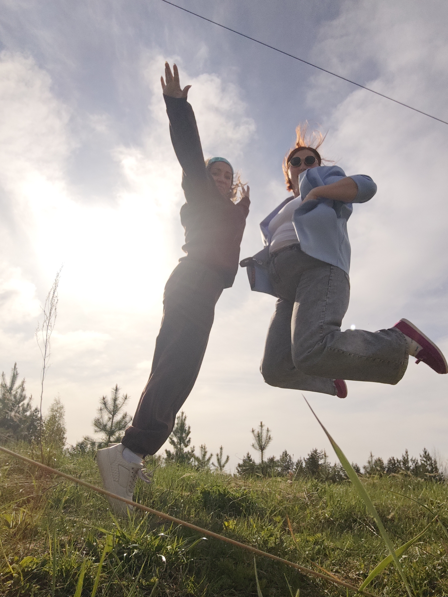 Two women are captured mid-air, jumping in a grassy field. The woman on the left wears a dark-colored jacket and pants with white sneakers. Her left arm is raised above her head with her hand open. The woman on the right wears a light blue blazer over a white top with gray jeans and pink sneakers. She is wearing sunglasses. The background shows a slightly overcast sky with some visible trees. The location is most likely in Chaikovsky, Russia.