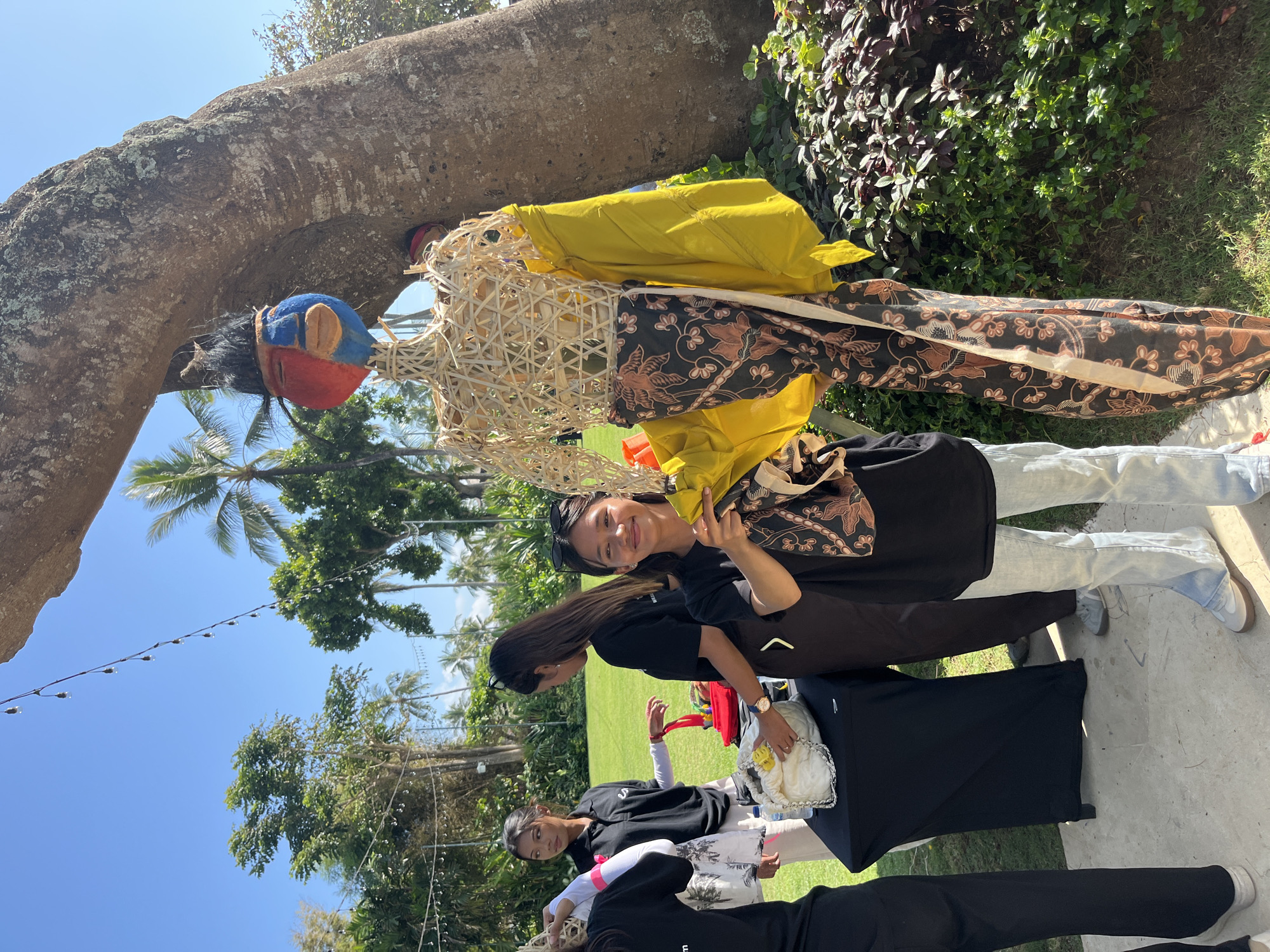 In Kuta Selatan, Indonesia, on a bright, sunny day, a group of young women are gathered outdoors near a large tree. One young woman, with dark hair and wearing a black t-shirt and light-wash jeans, stands prominently in the foreground, smiling at the camera. She is holding a patterned fabric bag. Beside her is a life-sized mannequin constructed from woven bamboo, adorned with a colorful, painted mask-like head featuring red, blue, and flesh tones, and black hair. The mannequin is dressed in a vibrant yellow garment draped over its shoulders and a long, dark fabric with a floral pattern. Other young women, dressed in black t-shirts, are visible in the background, some interacting with a black table draped with a white cloth and carrying bags. The setting appears to be a grassy area with lush green foliage and palm trees visible in the distance. String lights are hung in the upper portion of the frame, suggesting an outdoor event or gathering.