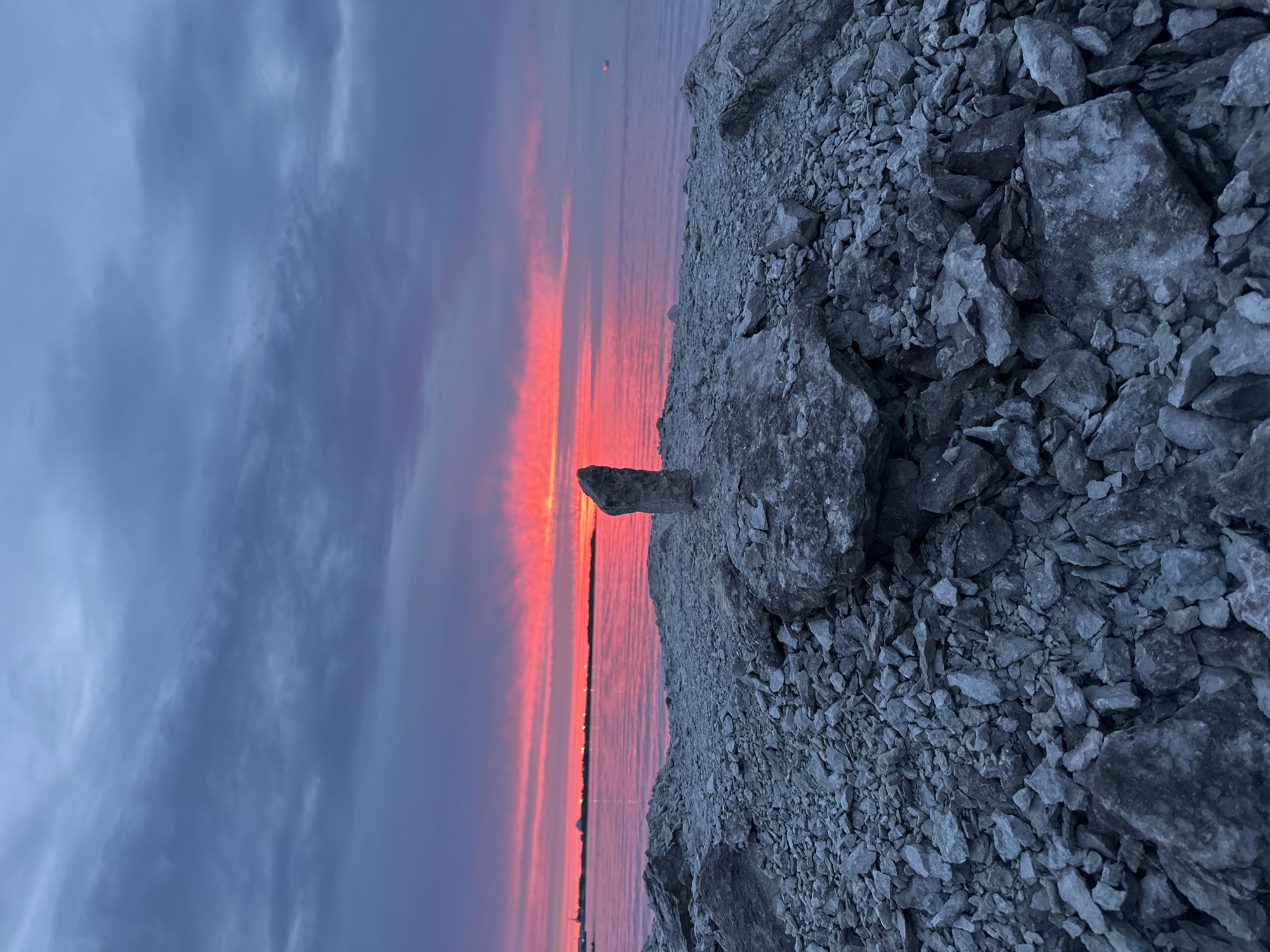 This image captures a dramatic sunset over the Baltic Sea, viewed from a rocky shoreline. The sky is ablaze with vibrant pinks and oranges, contrasting with the deep blues and purples of the clouds. The water reflects the colors of the sky, creating a shimmering effect. In the foreground, a precarious stack of stones stands as a solitary focal point. The shoreline itself is composed of rough, grey rocks and pebbles. In the distance, along the horizon, the faint outline of Tallinn's cityscape is visible, with lights twinkling along the coast, suggesting an urban setting. The overall atmosphere is serene and awe-inspiring, dominated by the natural beauty of the sunset.