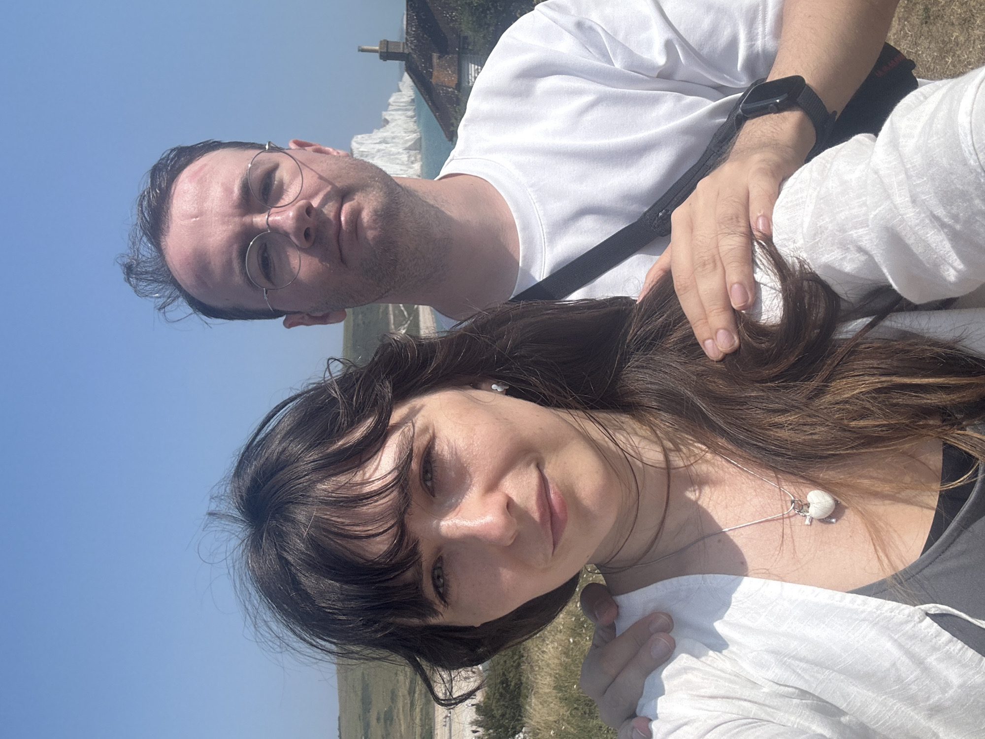 A selfie taken outdoors shows a man and a woman standing close together with a scenic backdrop. The woman is in the foreground, facing the camera and smiling slightly. She has dark, long hair that is partially covering her face and is wearing a white top and a necklace with a heart pendant. The man is behind her, also looking at the camera, wearing glasses and a white t-shirt. His arm is around her shoulder. In the background, there are rolling green hills, a body of water, and white cliffs, consistent with the location of Seaford, United Kingdom, specifically near the Seven Sisters chalk cliffs. The sky is clear and blue.