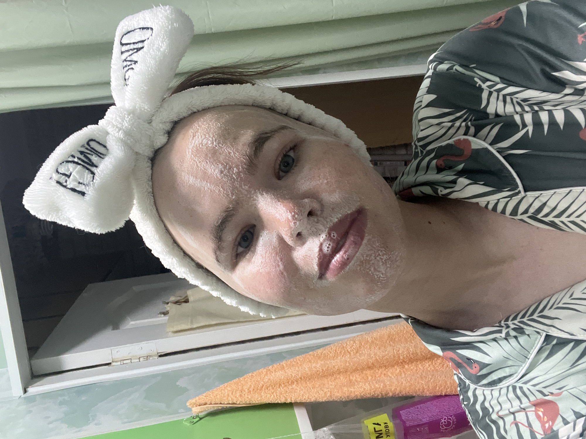 The image depicts a young adult female taking a selfie in what appears to be an indoor setting, likely a bathroom. She is looking directly at the camera with a neutral expression. Her face is covered in a white, foamy substance, consistent with a facial mask or cleanser, visible across her forehead, nose, cheeks, and around her mouth, where small bubbles are present on her lips. She has light-colored eyes and her hair is pulled back by a white, fluffy headband featuring two bow-like structures, each embroidered with the word "OMG" in black.
She is wearing a dark green (or teal/sage) pajama-style top with a white trim, patterned with white palm leaves and pink flamingos.
The background on the left features a wall with a green base color partially covered by a lighter, possibly textured or painted, light blue/teal pattern. An orange/peach colored towel hangs on a green hook mounted on this wall. Below the towel, partially obscured, is a purple container and a clear plastic bag with Cyrillic text, including "ДЛЯ" and "УЛОВК". A white door is slightly ajar in the middle-left background, revealing a darker space beyond and showing a visible metal hinge on its frame. On the right side of the image, a light green or mint-colored curtain is visible. The lighting suggests an indoor, possibly artificial, source. There are no visual cues within the image to confirm the specific location as the city of Chaikovsky, Russia, though the Cyrillic text suggests a Russian-speaking context.