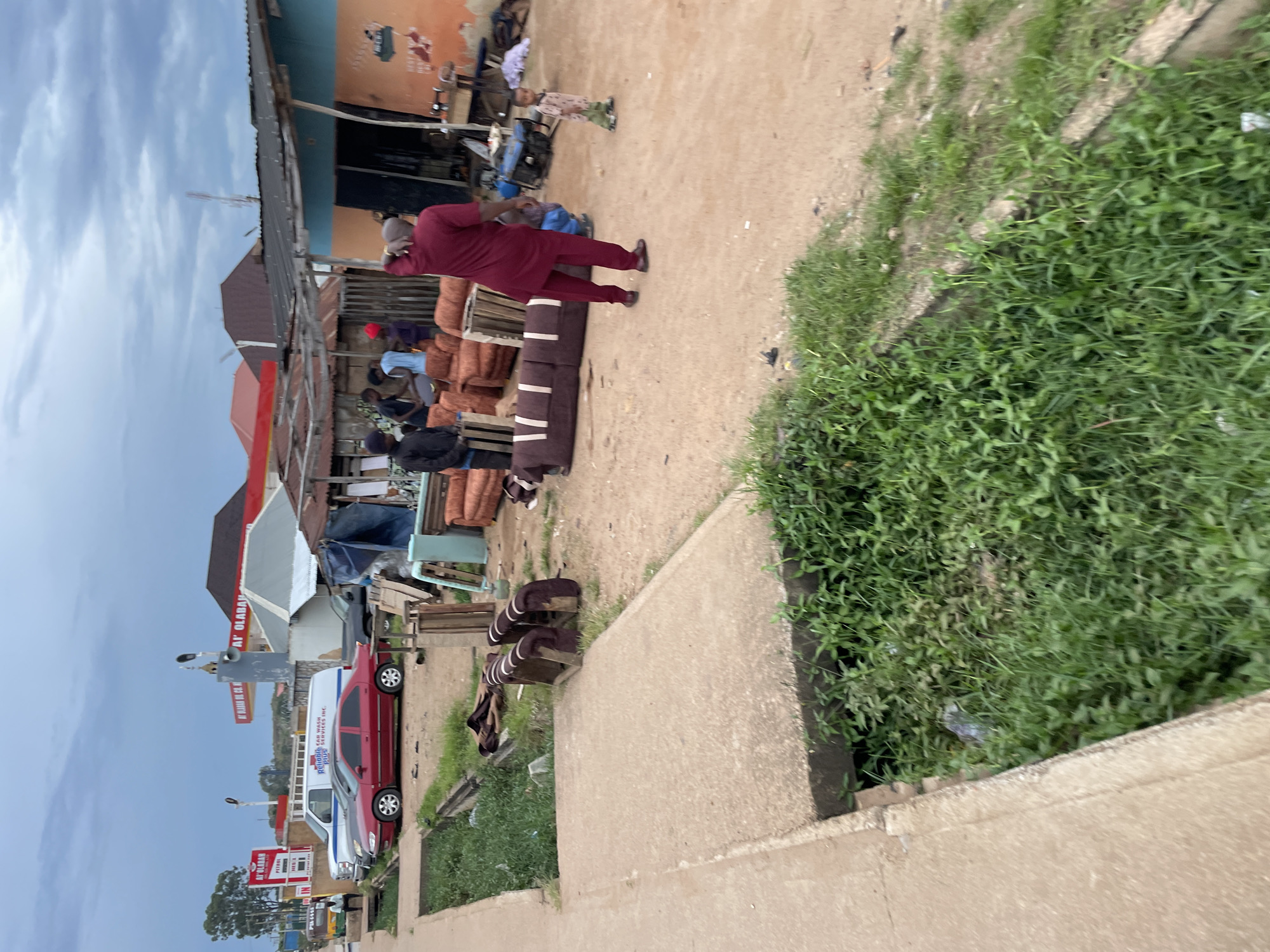 In Barakin Lerawi, Nigeria, the image depicts an outdoor scene with a focus on furniture and vehicles. A red sedan is parked on a dusty road, alongside a white van. In the background, a gas station with red and white signage is visible. Several people are gathered around furniture pieces, including what appears to be a long sofa and some individual seating units. The environment is characterized by a mix of structures, including what looks like a workshop with a corrugated metal roof, and an open area with exposed earth and patches of green grass and vegetation. A small child stands near the workshop. The sky is overcast.