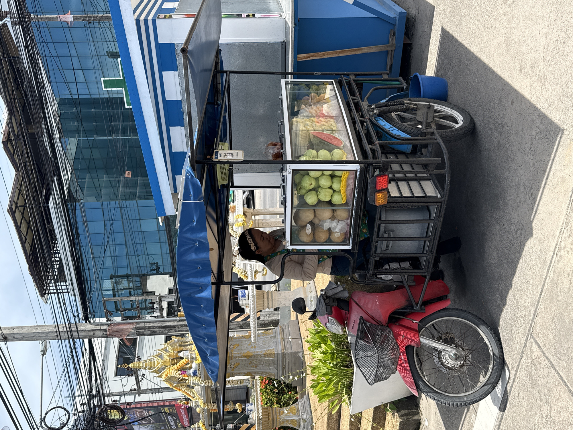 A street food cart, consisting of a red motorized tricycle with a canopy and a glass display case filled with fruit, is parked on a paved sidewalk in the city of Ko Samui District, Thailand. A woman is standing behind the cart, attending to the display. The cart is positioned in front of a modern building with blue glass windows and a golden temple structure. The environment appears to be a busy urban area, indicated by the overhead power lines and a billboard on the left.
