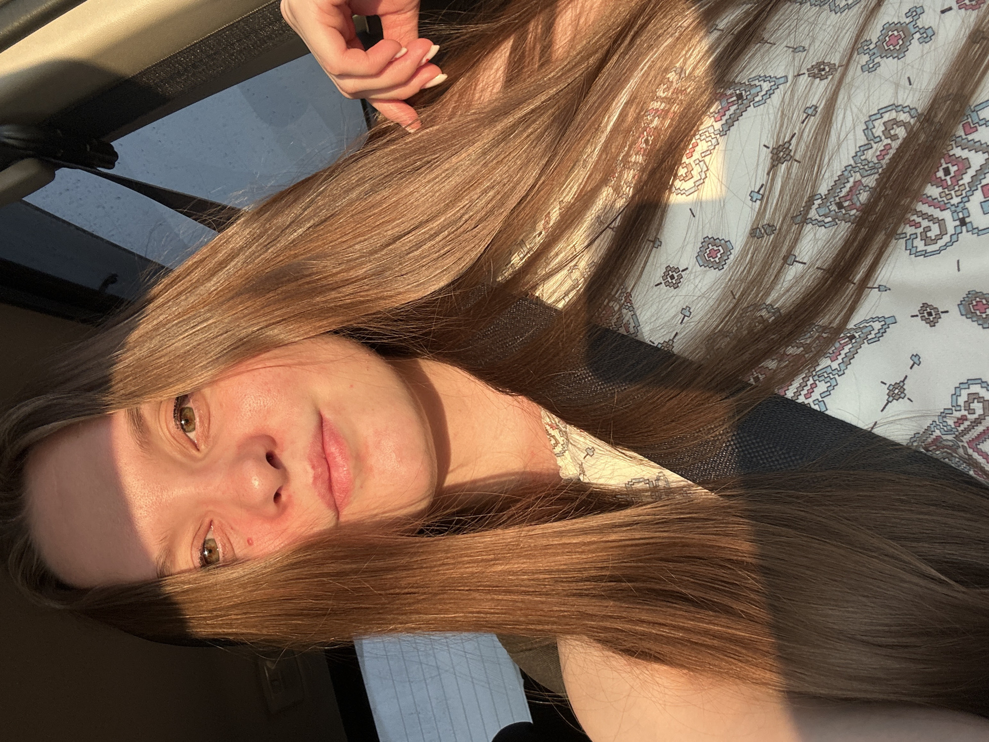 A young woman with long, auburn hair is captured in a selfie taken inside a vehicle, likely a car, as indicated by the seatbelt and window. She is wearing a light-colored, patterned top. The lighting suggests it's either early morning or late afternoon, with warm sunlight illuminating her face and hair. The background shows a car interior and a window with reflections, hinting at an outdoor environment. The woman's hand is visible near her hair, suggesting a casual pose. There is no specific indication of the location within the city of Chaikovsky, Russia, beyond the fact that it is an urban or suburban setting.