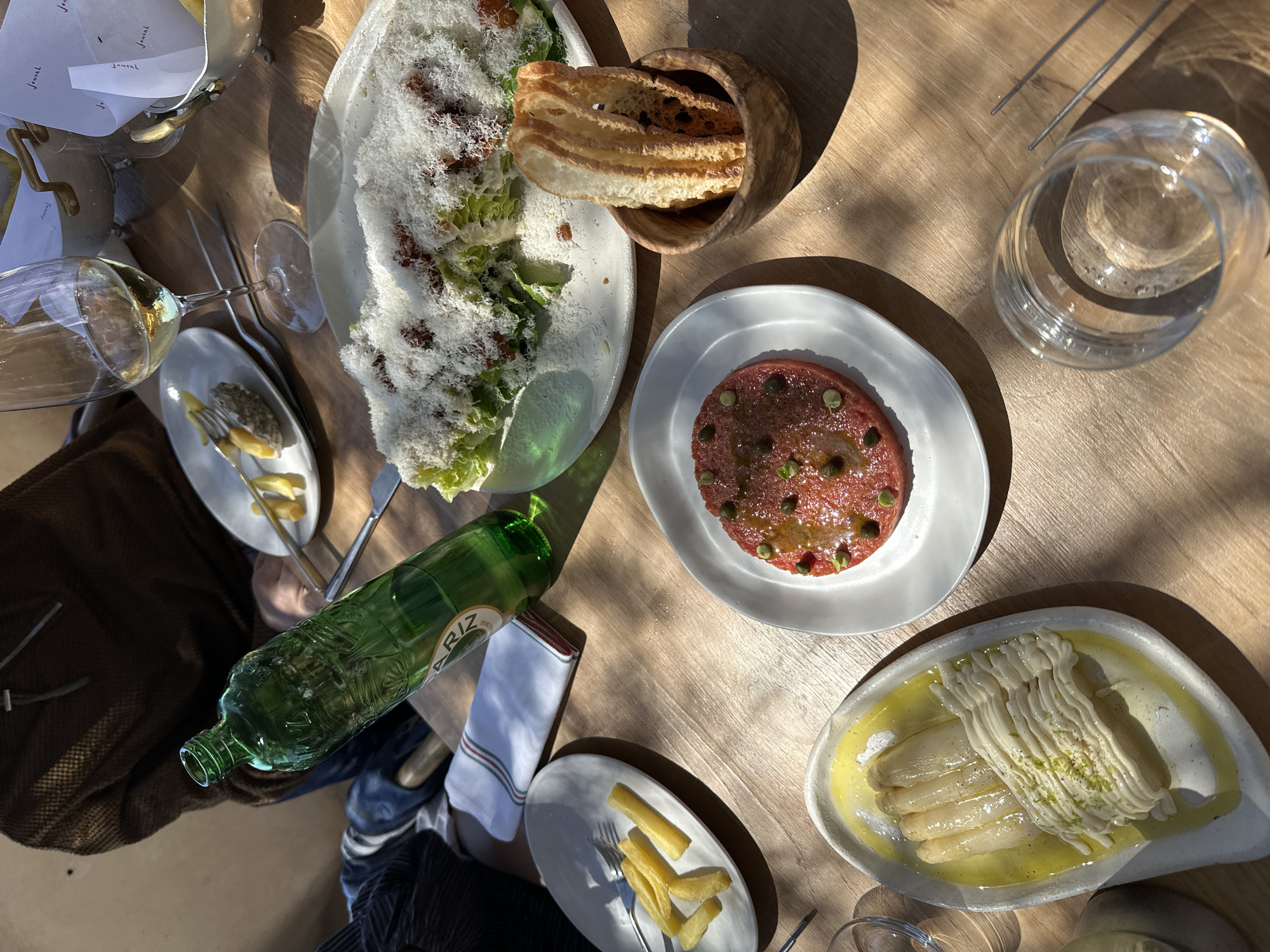 An overhead shot captures a variety of dishes on a wooden table, likely in a restaurant in Sant Josep de sa Talaia, Spain. A Caesar salad liberally sprinkled with grated cheese and bacon bits, accompanied by toasted bread in a wooden container, is present. A red steak tartare garnished with green capers and drizzle of olive oil sits on a white plate. A side dish of white asparagus with sauce is also visible. A green glass bottle, presumably of water, is on the table. There are multiple sets of silverware and glassware. Part of a person is visible to the left of the bottle, wearing a dark jacket and jeans. The lighting suggests a bright, sunny day.