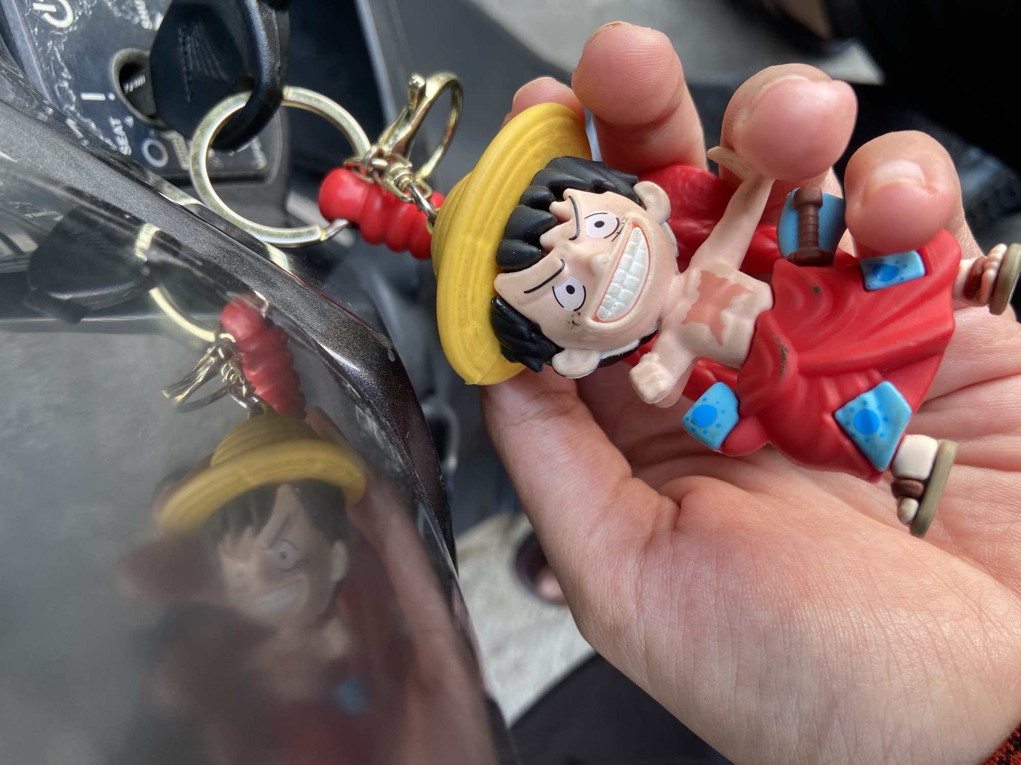 A close-up shot shows a person's hand holding a Monkey D. Luffy keychain. The keychain is a small, plastic figurine of the character from the anime series One Piece, wearing his signature straw hat and red outfit. The figurine has a wide grin and is detailed with black hair, white eyes with black pupils, and a scar on his chest. Red beads and a gold keyring are attached to the figurine. In the background, a motorcycle ignition is visible with a key inserted. The overall environment suggests an outdoor setting, possibly in a residential area or a parking lot.