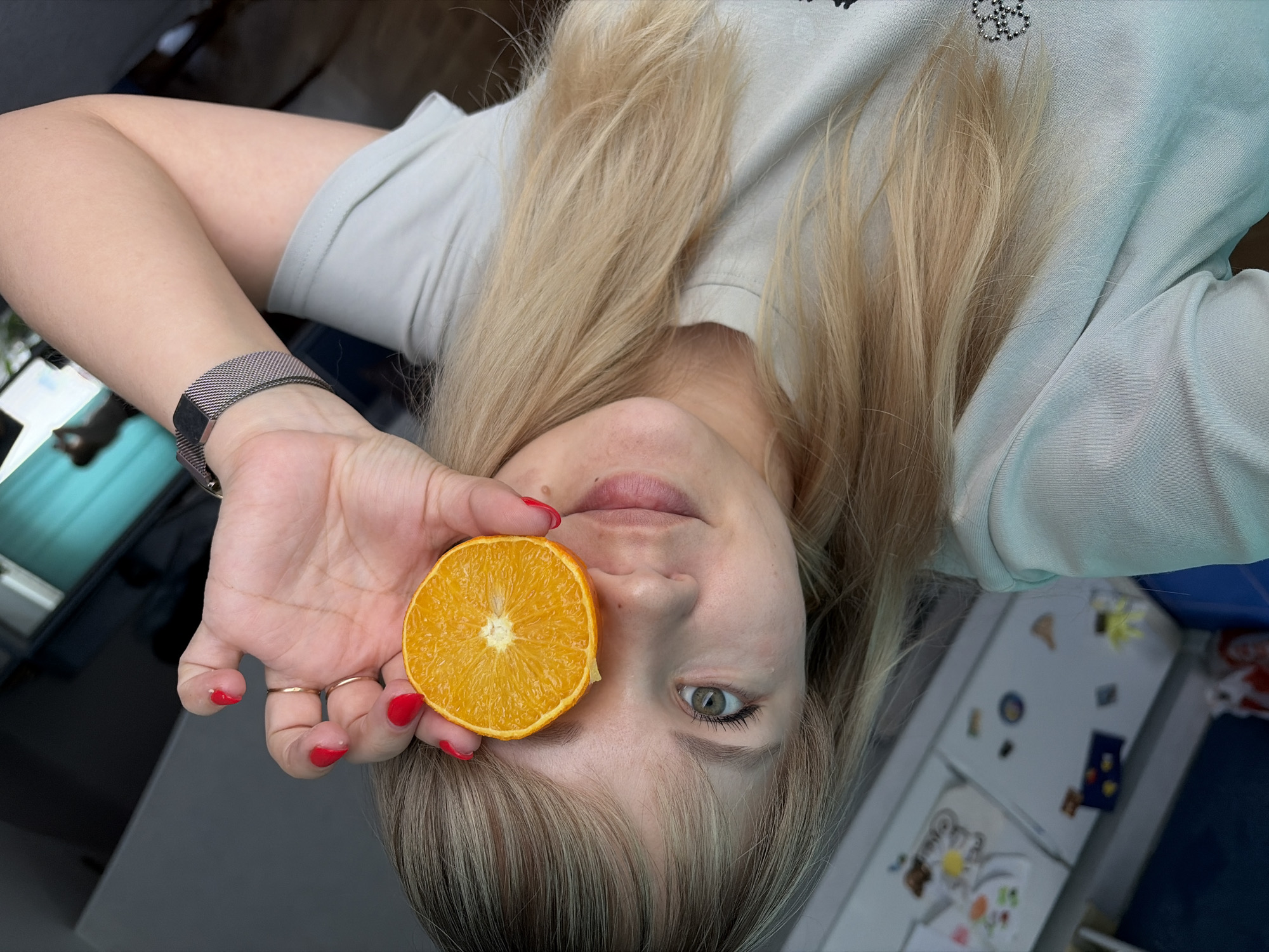 A young woman with long, blonde hair is holding a half-cut orange in front of her eye. She is wearing a light-colored t-shirt and has a silver watch on her left wrist. Her fingernails are painted red, and she wears two gold rings on her fingers. Behind her is a refrigerator with children's drawings on it. The location is likely in a domestic kitchen within the city of Chaikovsky, Russia.