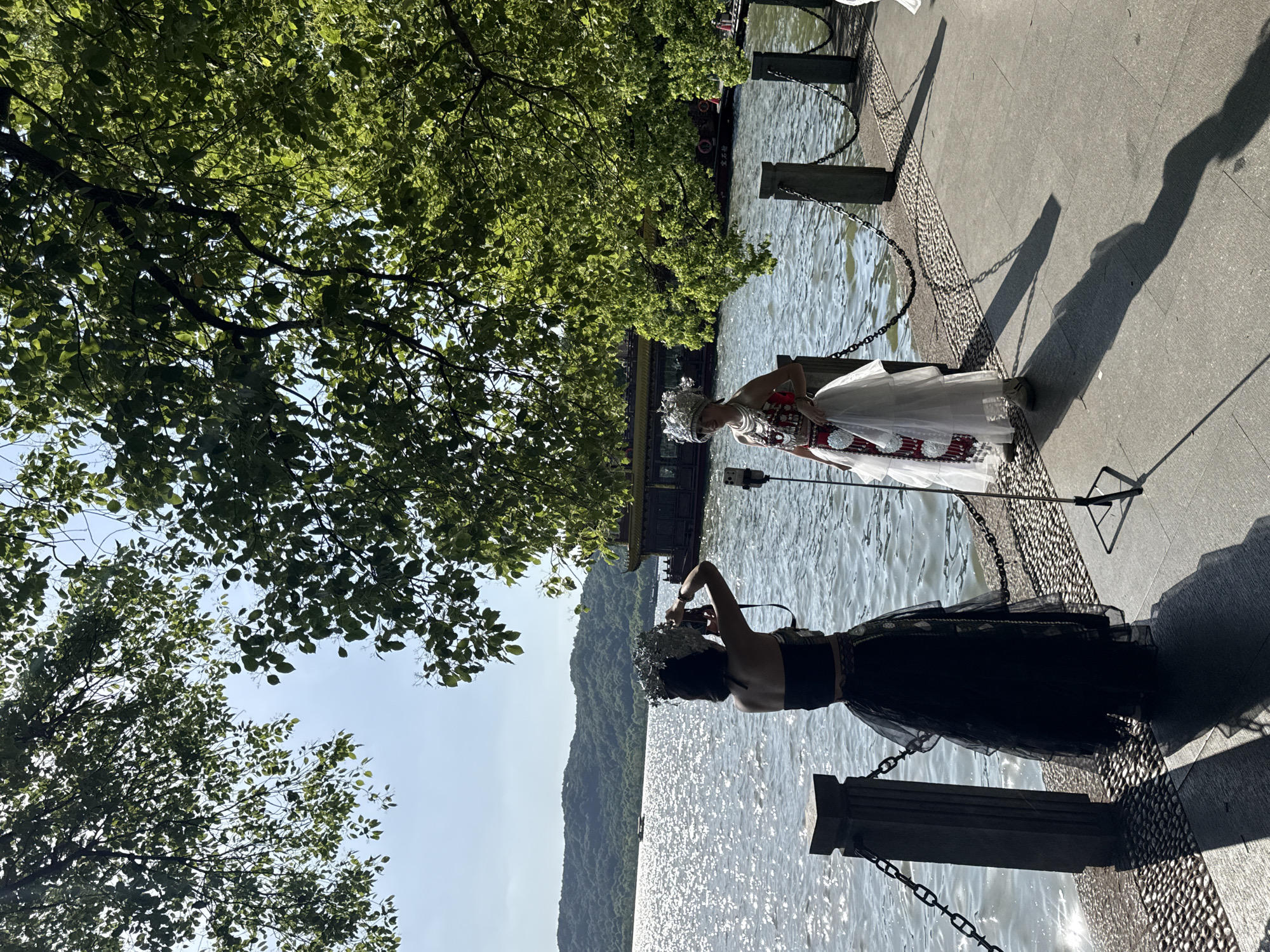 On a sunny day in Xihu District, China, two individuals are positioned near a body of water, possibly a lake or river, bordered by a paved walkway and chain-linked posts. The foreground features a person in a black dress holding up a camera, seemingly photographing another person dressed in a white traditional costume with ornate headwear. In the background, a lush, green hill rises, and a boat floats on the water. Above, a canopy of tree branches and leaves filters the sunlight. The scene suggests a photo shoot or a performance of some kind.