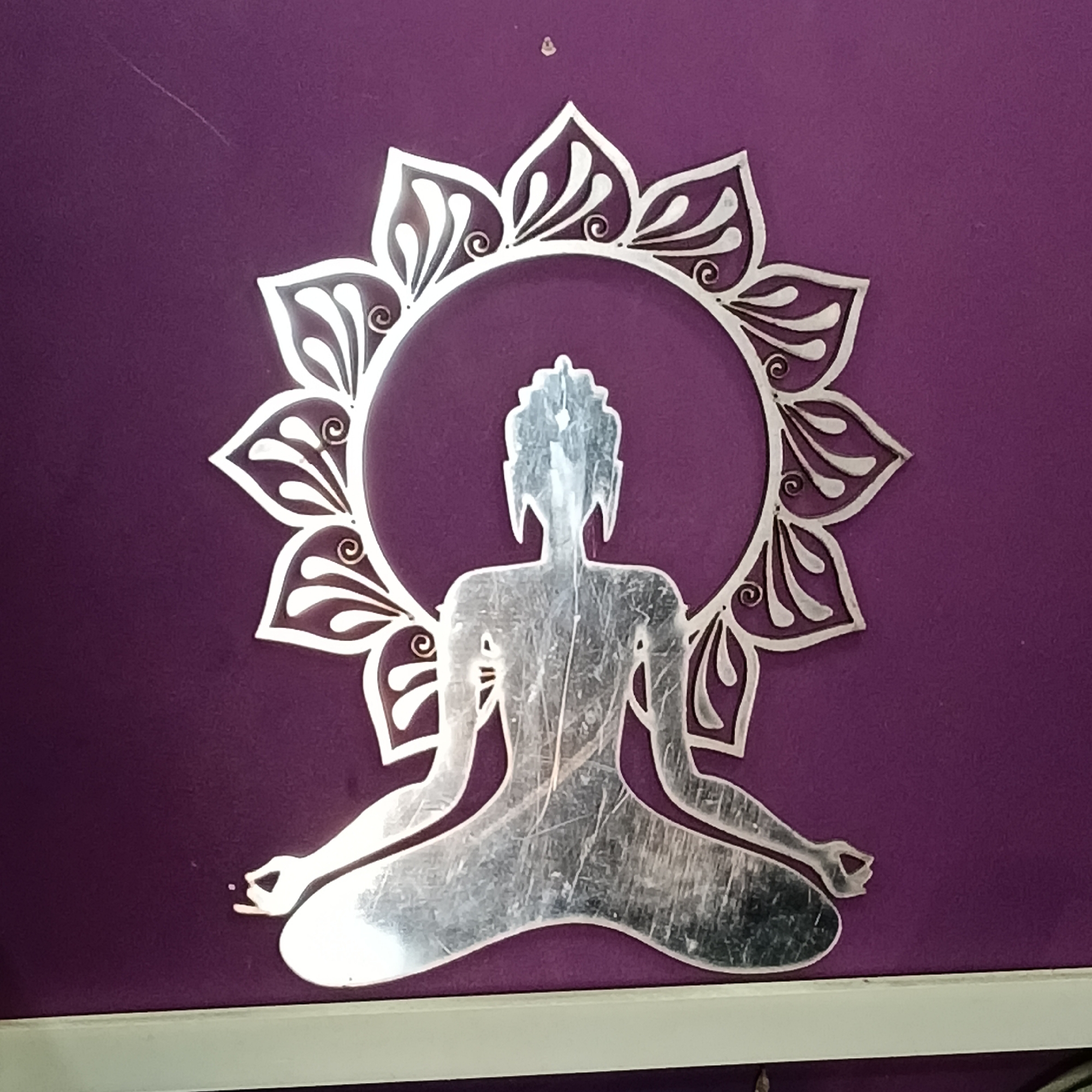A silver-colored metal artwork is affixed to a purple wall. The artwork depicts a stylized figure in a meditative pose, enclosed within a circular floral halo. The wall appears to be indoors, as suggested by the lighting. The composition consists of a figure in a meditative pose surrounded by a halo of lotus petals. The artwork is metallic, while the background is a solid purple. There is no visible evidence to confirm that the location is in Oxel, India.