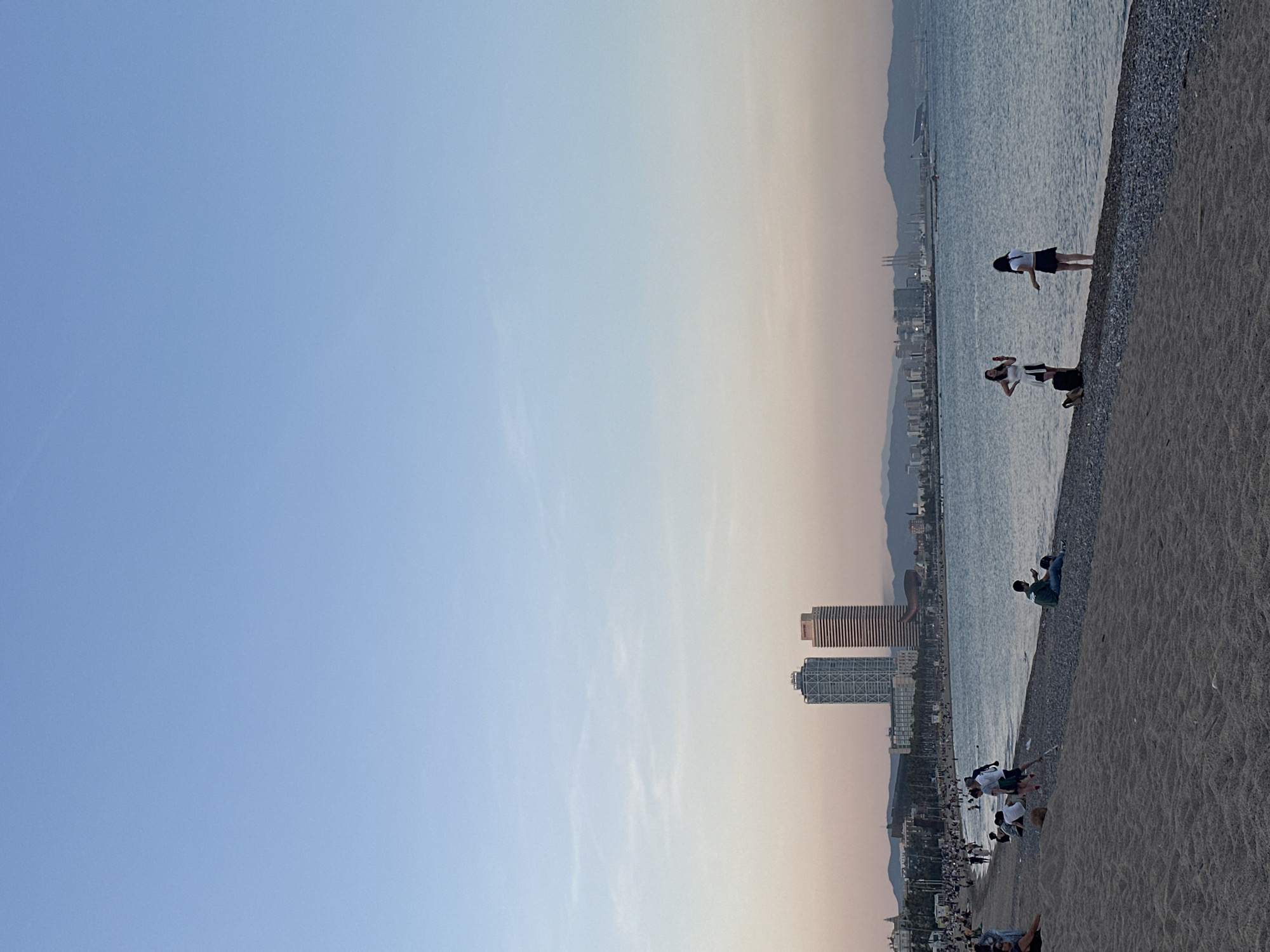 The image captures a coastal scene in Barcelona, Spain, with a beach extending along the right side of the frame. The sky transitions from a blue hue to an orange tint towards the horizon. In the distance, the city skyline is visible, including prominent buildings such as the Hotel Arts and the Mapfre Tower. People are scattered along the beach, some standing near the water's edge, others sitting or lying on the sand. The sea appears calm.