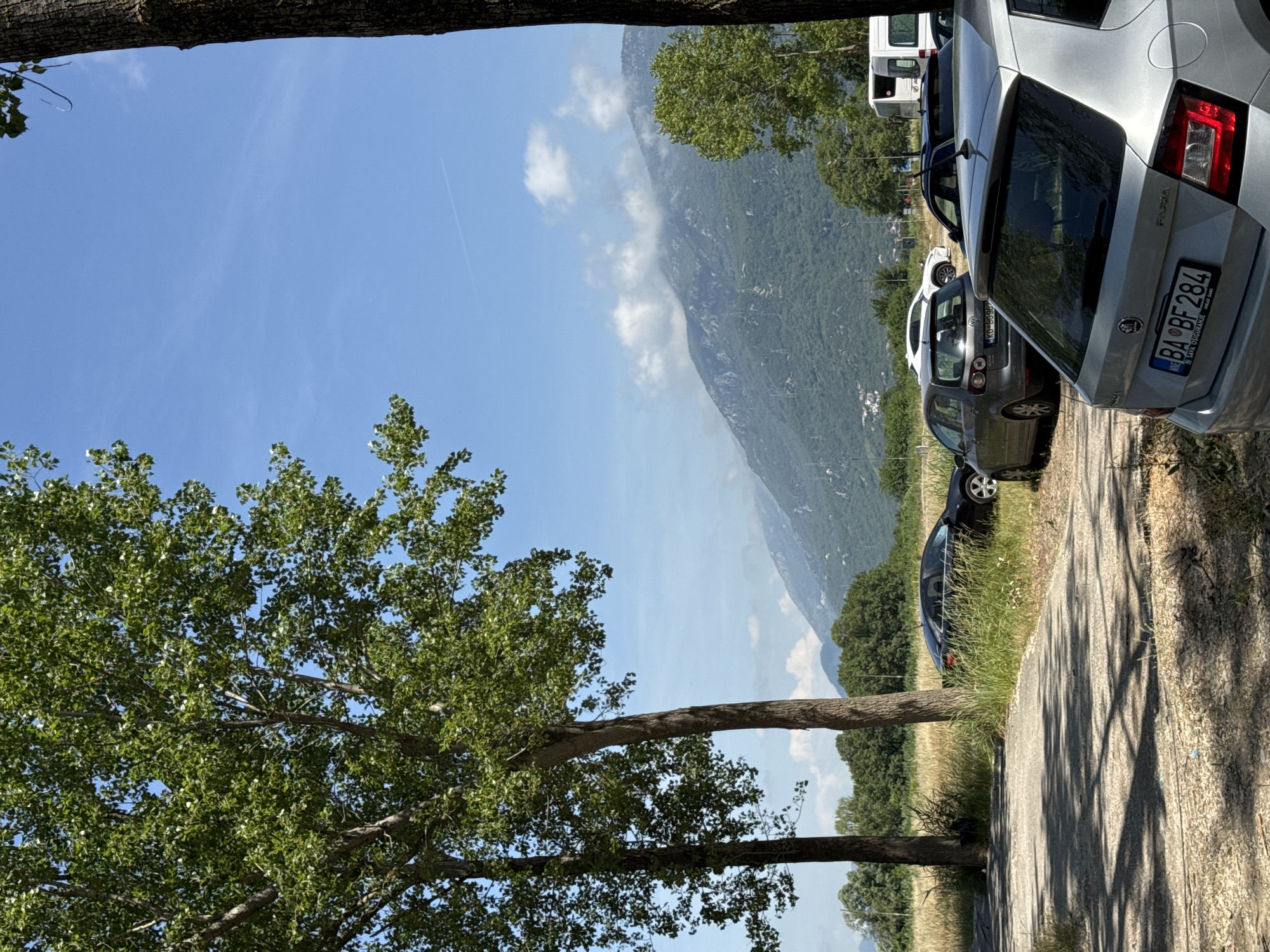 The image depicts a roadside scene near Prijevor, Montenegro. A line of parked cars, including a silver Skoda Fabia with the license plate "BA BF 284," is visible along the right side of the frame. The road itself is paved, with grassy verges bordering it. A mountain rises in the background, partially obscured by clouds. Trees frame the left side of the image, with their foliage partially blocking the sky. The sky is blue with scattered clouds, and a contrail is visible. The season appears to be summer, based on the green foliage and sunny conditions.