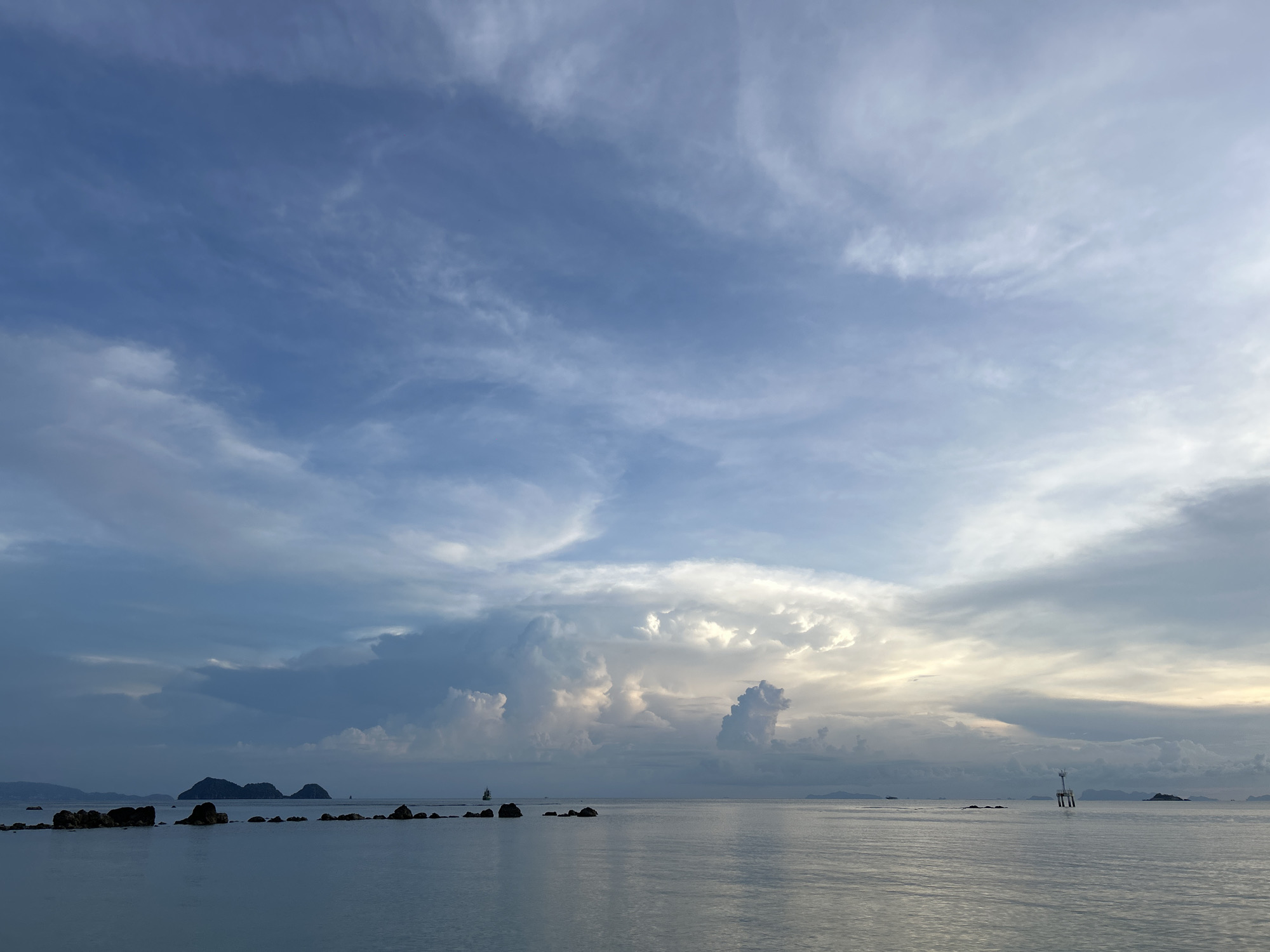 The image shows a body of water with several rocky islets and larger islands in the distance. The sky is filled with clouds of varying density and color, ranging from deep blues to bright whites, suggesting a dynamic weather pattern or possibly a sunrise or sunset. A navigational marker stands in the water on the right side of the frame. The location is Baan Sri Thanu, Thailand.
