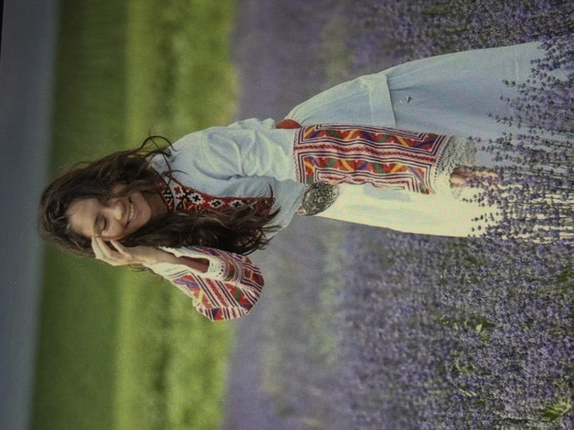 A young woman with long, dark, wavy hair is standing in a field of purple flowers. She is wearing a light blue dress with embroidered sleeves and a red belt. The embroidery on the sleeves is in a pattern of red, white, green, and yellow. Her right hand is raised to her hair, and she is smiling. The background is a blurred green field with a dark sky above. The overall impression is one of peace and happiness.