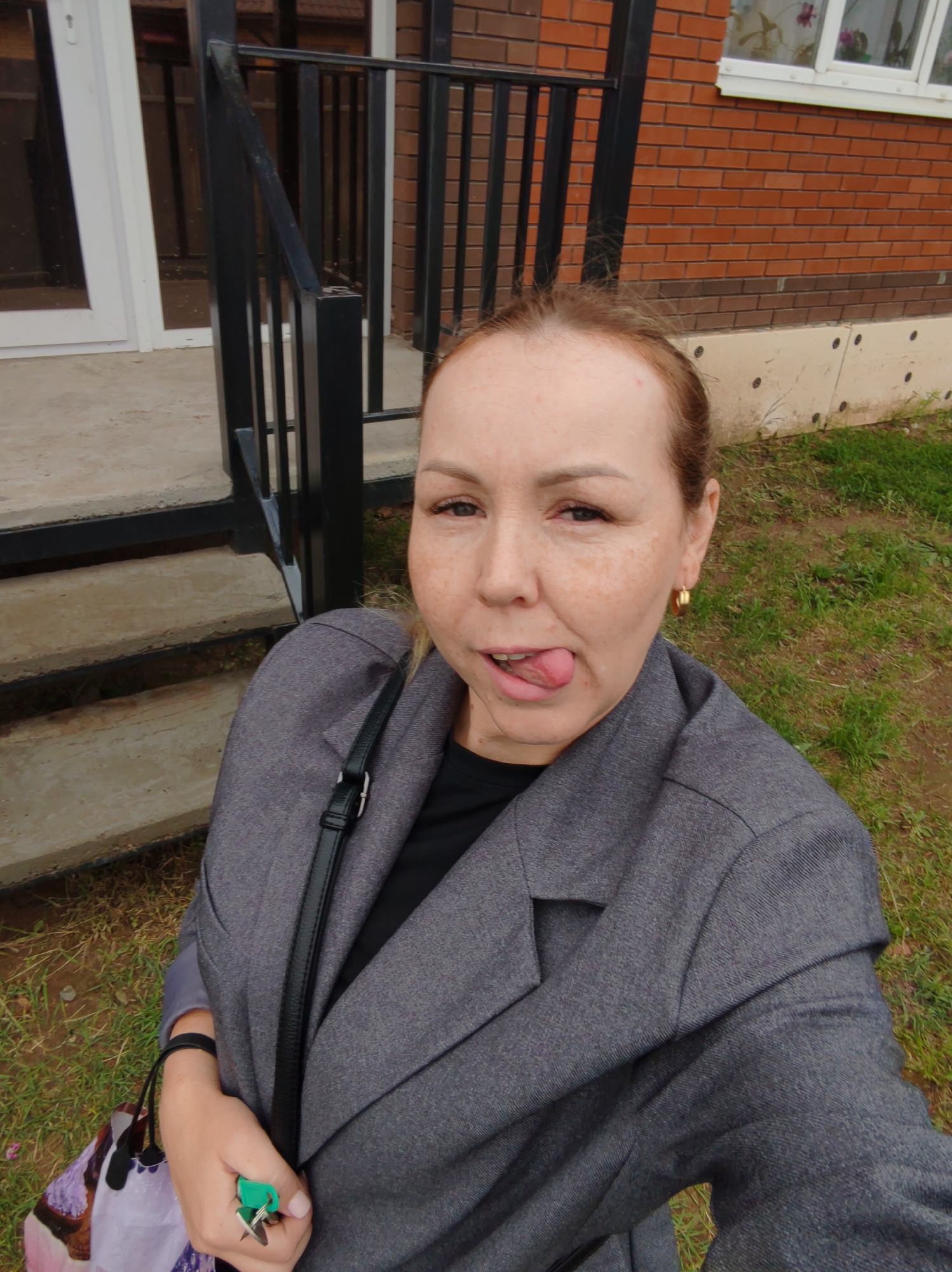A woman is seen outdoors in the city of Chaikovsky, Russia. She has fair skin, freckles, and brown hair pulled back. She is wearing a gray blazer over a black top. She has a black purse strap across her torso and holds keys in her left hand. She is sticking her tongue out. Behind her is a brick building with a black metal staircase leading to a white door. There is grass on the ground.