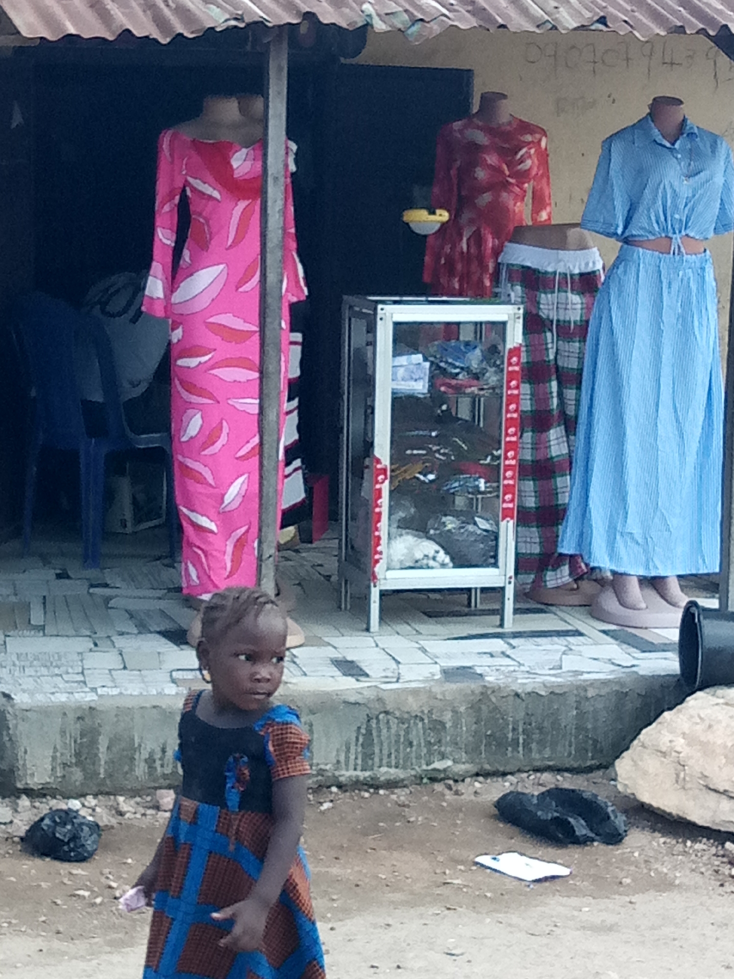 A young girl stands in the foreground of the image, facing towards the right. She has dark skin and her hair is styled in small braids. She is wearing a dress with a blue bodice and a patterned skirt in brown and blue. To her left, a black garbage bag is visible on the ground. Behind her, a stall is set up, displaying clothing on mannequins. One mannequin on the left wears a long, pink dress with a leaf pattern. In the center, a display case is filled with various clothing items. To the right, two mannequins are dressed in a red patterned top and a blue striped shirt with a matching blue pleated skirt. The stall has a corrugated metal roof and the ground is paved with light-colored tiles. The background appears to be an outdoor setting with a wall displaying a phone number.