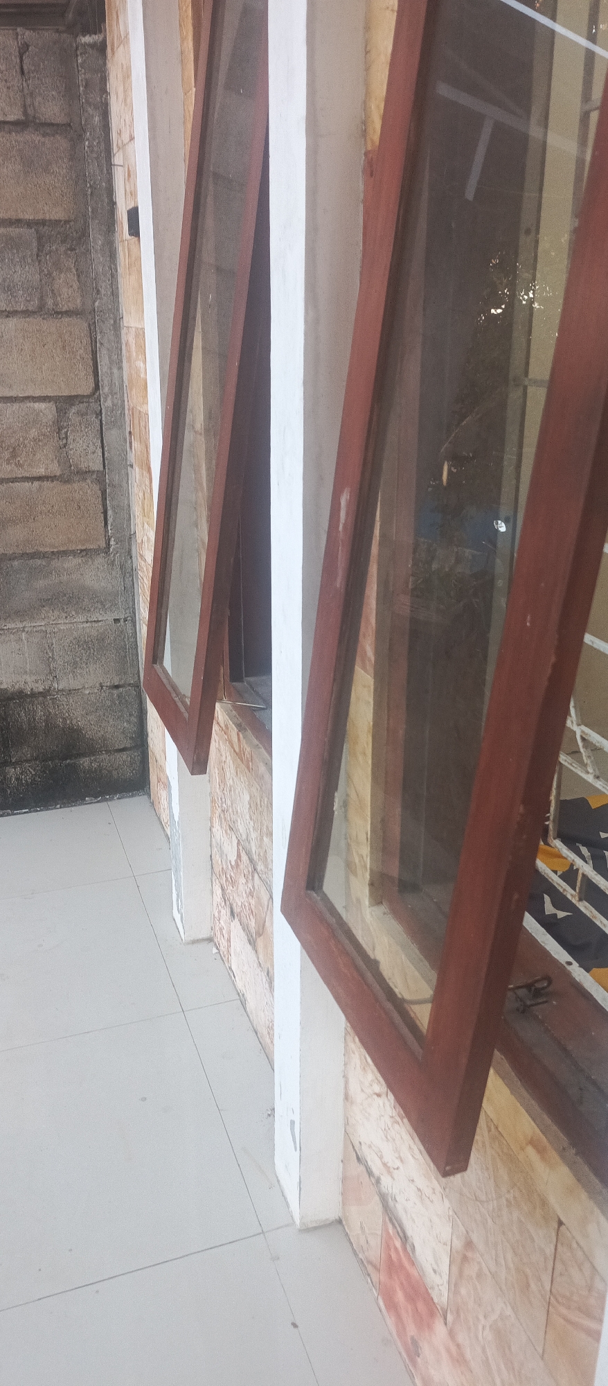 This image depicts an outdoor scene in Jatisari, Indonesia, showing an exterior wall with two open windows. The windows have dark brown wooden frames and glass panes. One window is open at an angle, revealing the interior which is dimly lit and appears to be a room. The wall is constructed with rough-hewn stone blocks on the left and lighter-colored, textured stone tiles on the right. A white pillar is visible between the two windows. The ground in front of the windows is paved with light-colored square tiles. The overall environment suggests an architectural detail of a building, possibly a house or a commercial establishment.
