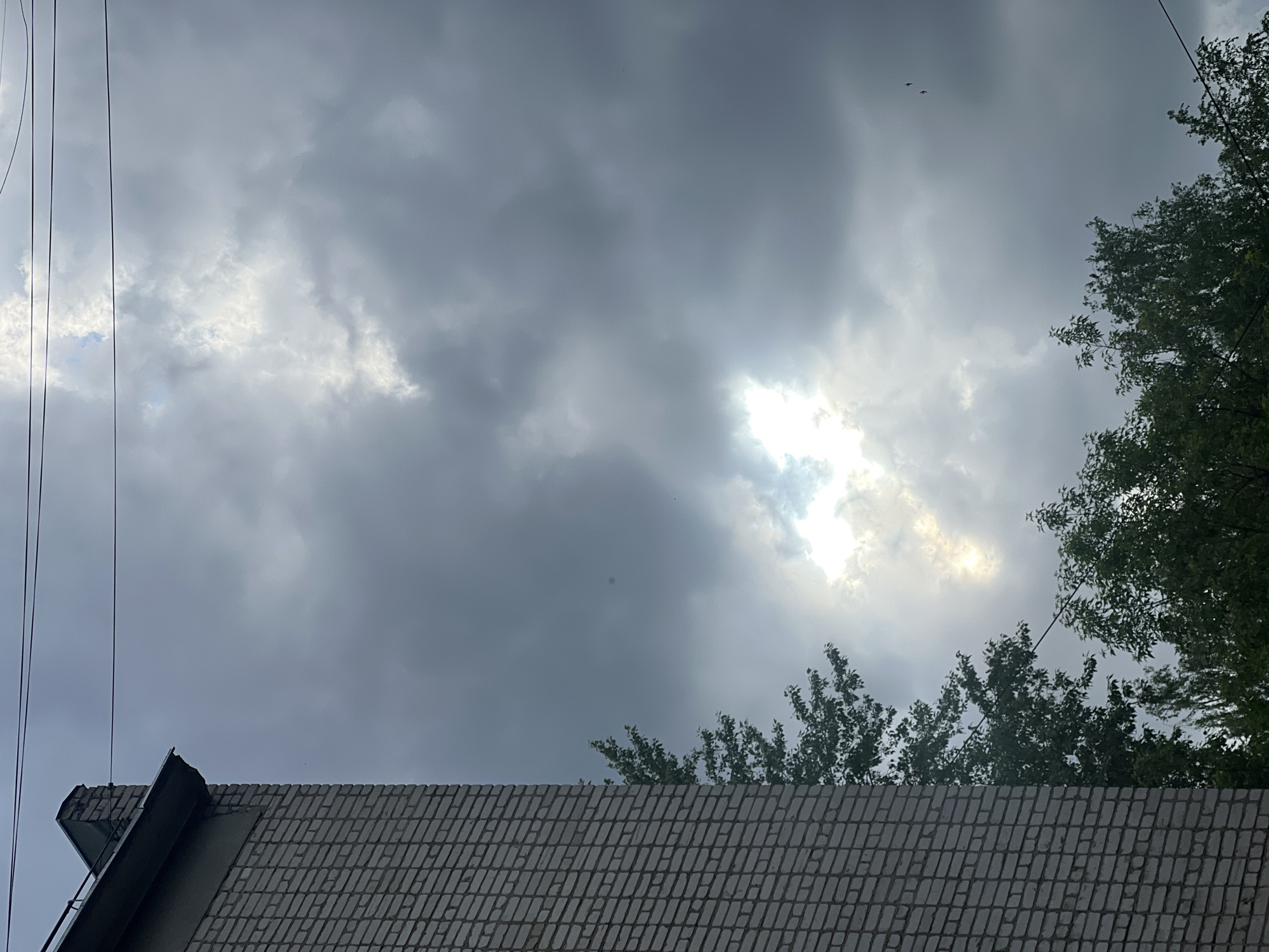 The image shows a view looking upwards from a ground-level perspective. In the foreground, there's a grey tiled roof, partially obscuring the scene. Above the roof, there are trees visible on the right side of the frame, and a few branches are visible above the roof. The sky is filled with dark, overcast clouds, with a patch of bright light piercing through them. Electrical wires run across the upper-left side of the frame. The location is the city of Chaikovsky, Russia.
