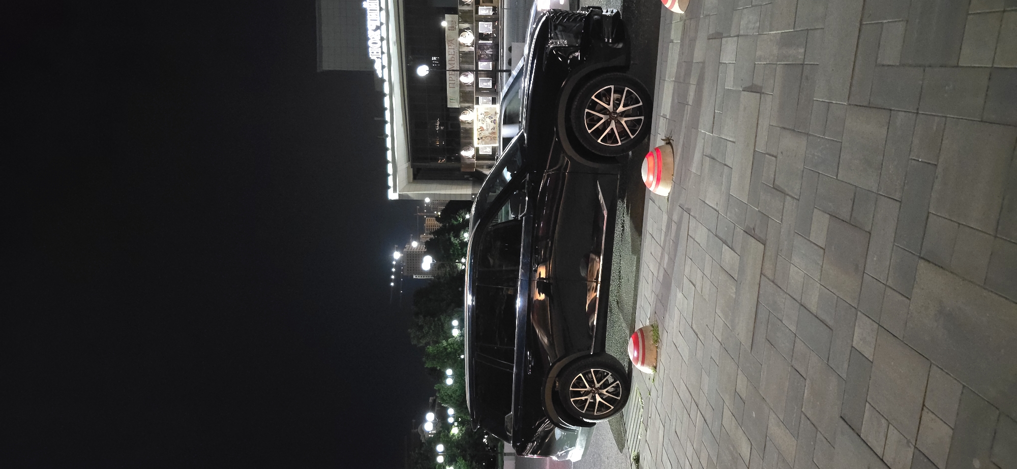 The image depicts a dark-colored SUV parked on a paved sidewalk in Grozny, Russia, at night. The vehicle is positioned next to several low, red and white striped bollards. In the background, buildings are illuminated, with one structure appearing to be a theater or performance venue based on visible signage. Streetlights are visible along the road, illuminating trees and urban structures. The overall scene is a typical urban nightscape.