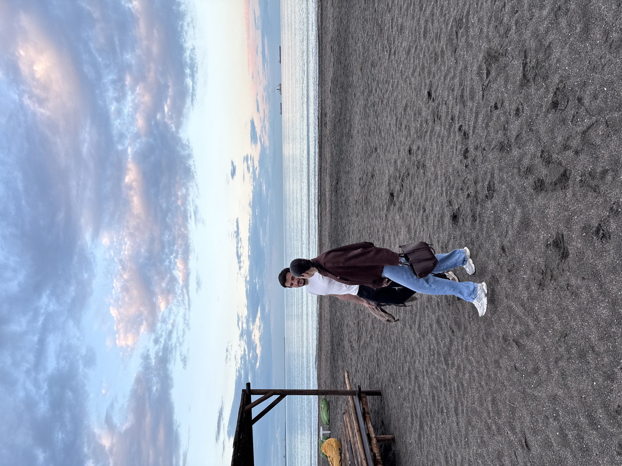 Two people stand on a beach with dark sand in Burgas, Bulgaria. They are turned sideways to the camera, with one person in a brown jacket and jeans, and the other in a white shirt. The sea is in the background, with a few ships visible on the horizon. The sky is filled with clouds, hinting at sunset. A wooden structure is visible in the lower left corner.