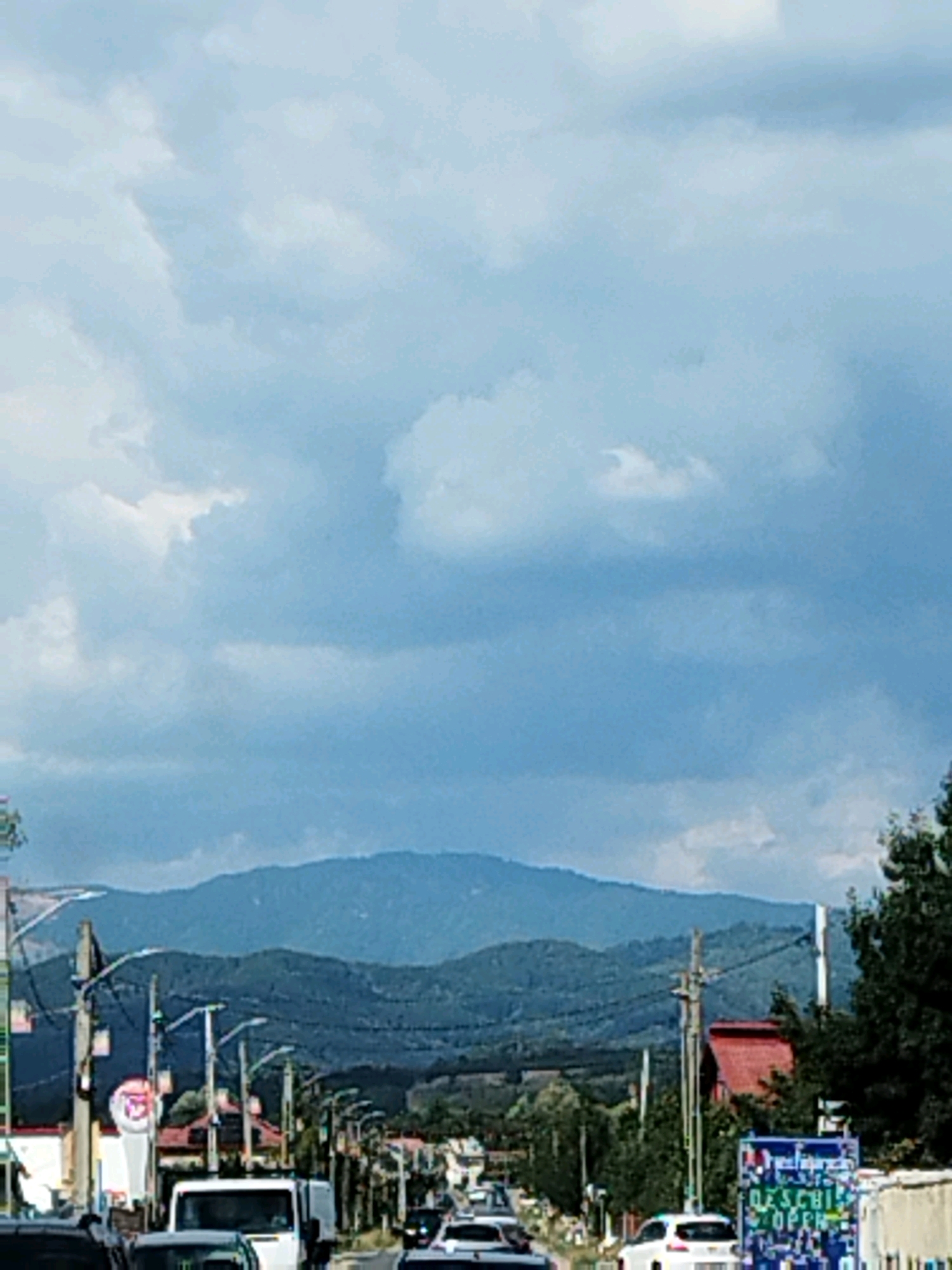 The image captures a view of a road in Albeștii Ungureni, Romania, with mountains in the background. The sky is filled with large, fluffy clouds, casting shadows on the landscape below. A road stretches into the distance, lined with trees and buildings, including houses with red roofs and what appears to be a shop or service station with signage. Several cars are visible on the road, suggesting it is a busy thoroughfare. Electrical poles and wires are a prominent feature, typical of a rural or semi-urban setting. The overall impression is that of a daytime scene in a town or village located near a mountainous region.