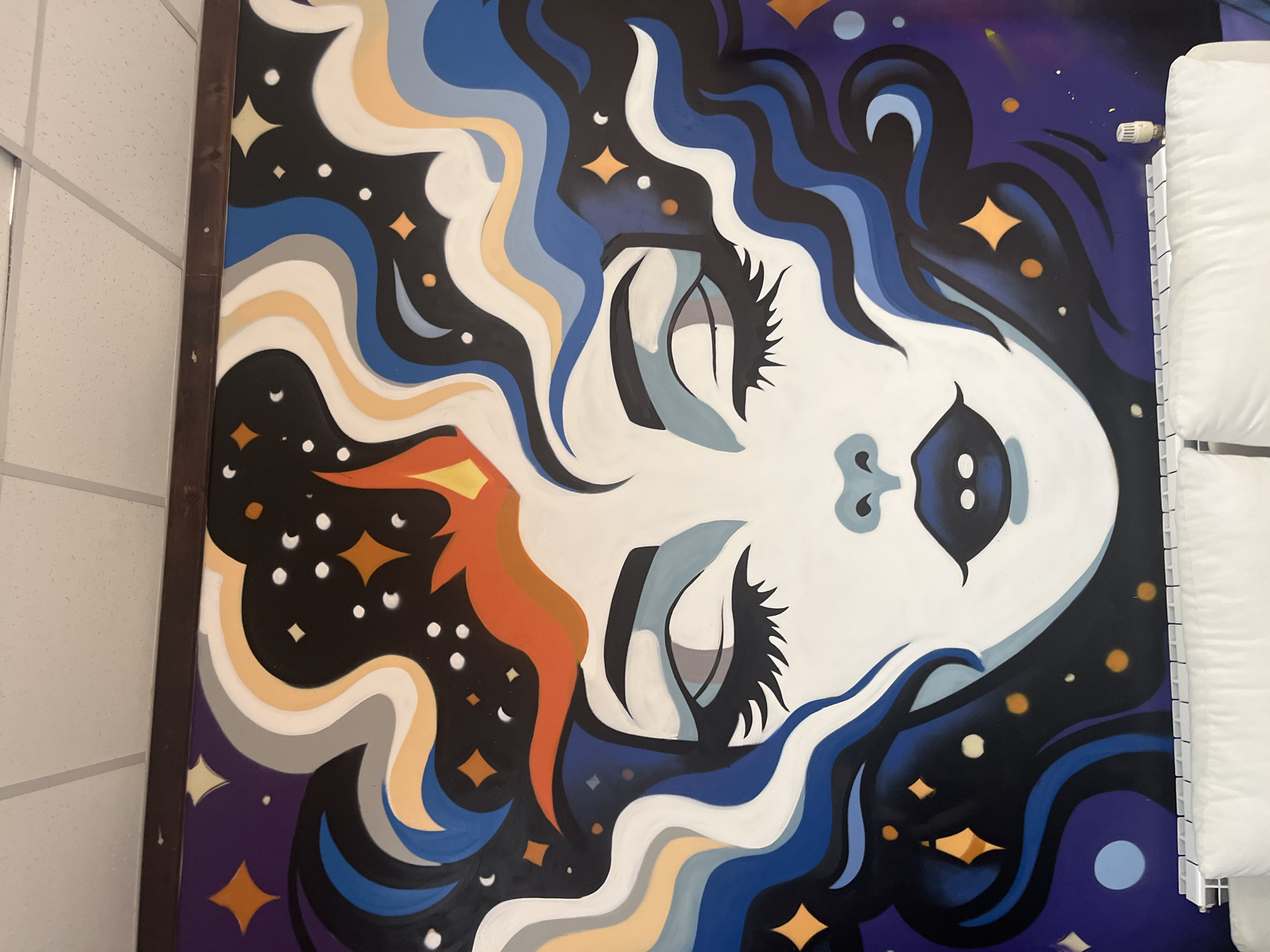 The image shows a large mural painted on a wall. The mural depicts a stylized face of a woman with closed eyes, her hair flowing outwards like waves and stars. The face is predominantly white with blue lips and light blue accents around the eyes and nose. Her hair is a mix of dark blue, black, white, orange, and gray wavy lines, interspersed with yellow stars and white dots representing stars or cosmic dust. The background is a dark purple and black gradient, further enhanced by various celestial elements. The mural is mounted above a white radiator and a white couch. The ceiling above the mural is a standard white suspended ceiling with visible tiles and metal grid. The mural is likely located indoors, possibly in a commercial or public space, given the nature of the artwork and the visible radiator and ceiling. There is no discernible information in the image to pinpoint its location to the city of Vratsa, Bulgaria.
